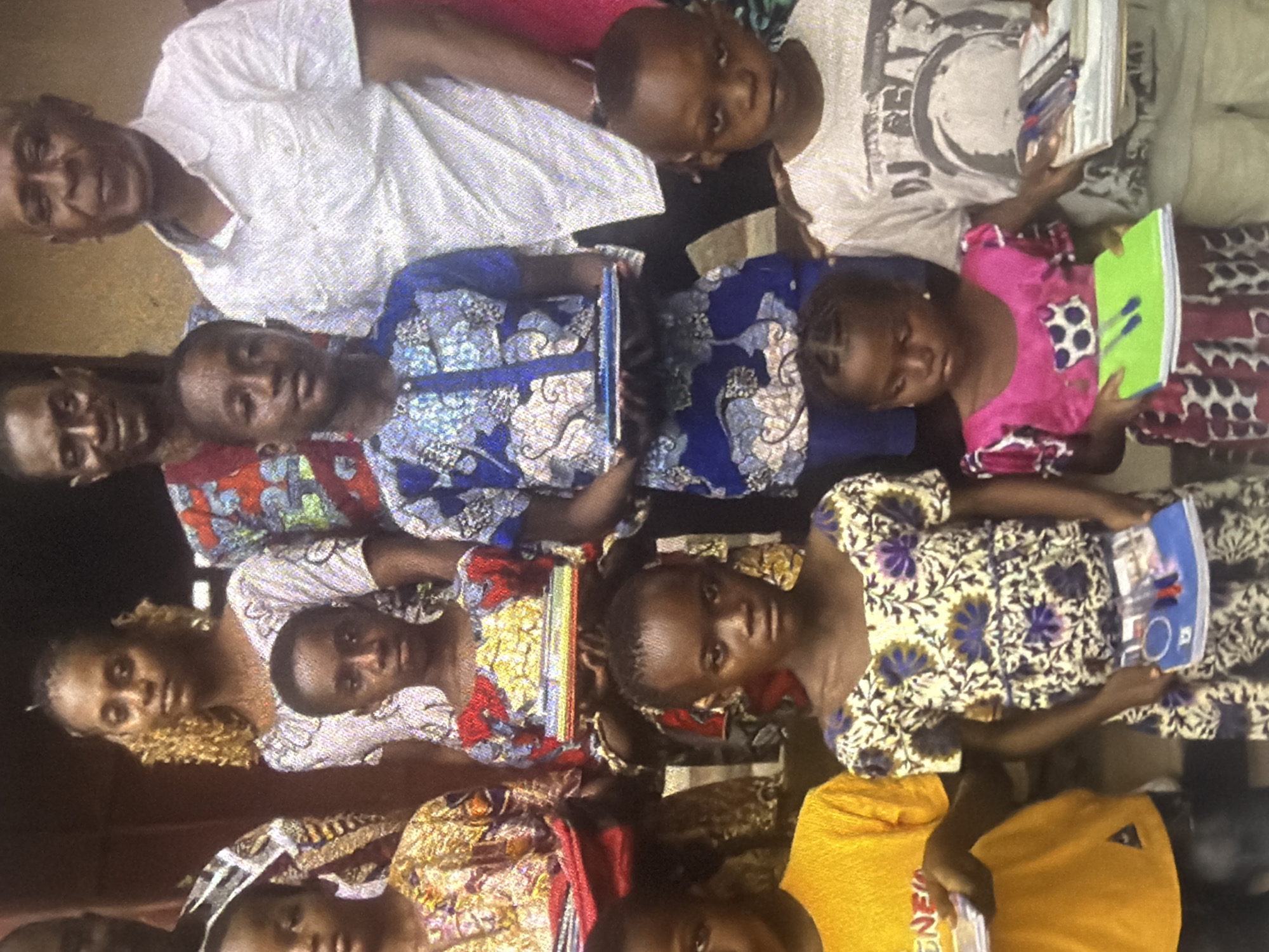 The image depicts a group of people, primarily children and some adults, gathered together. They are all looking towards the camera, suggesting a posed photograph. Many of the individuals are wearing colorful, patterned clothing, indicative of a cultural setting. Some of the children are holding books or notebooks, implying an educational context or a moment of study. The scene appears to be indoors or in a sheltered outdoor area, with a rustic or simple background. The lighting suggests it could be daytime, though the exact time of day is not discernible. The overall mood seems to be one of community and engagement. One child is holding a bright green notebook with pens, while another child is holding an open book with images. There is no visible text that can be clearly deciphered.