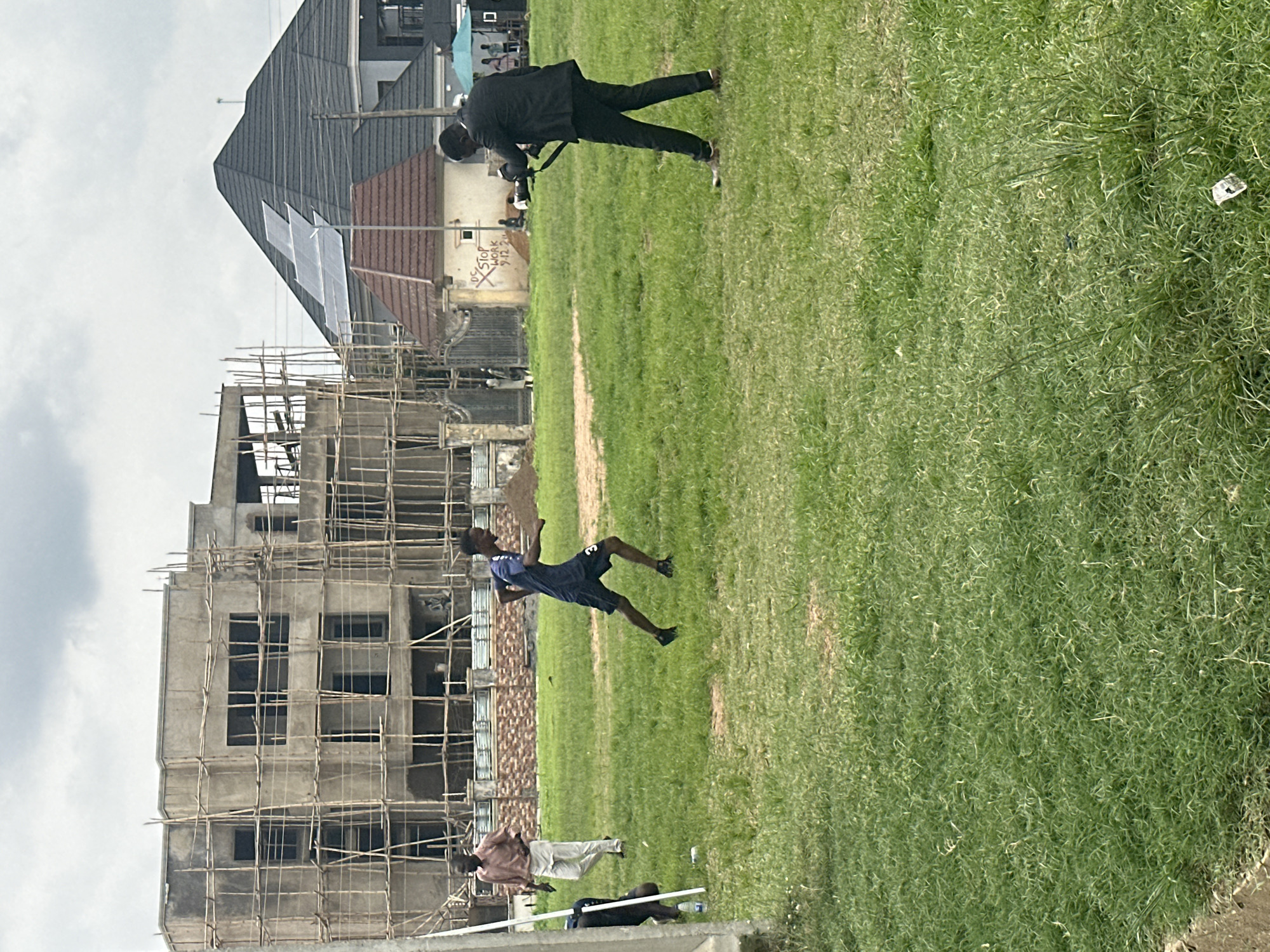 The image depicts an outdoor scene in what appears to be Abuja, Nigeria. The primary subjects are a group of people engaged in various activities on a grassy field. In the foreground, a construction site is visible, with a multi-story building under construction, surrounded by scaffolding made of bamboo. The building's concrete structure and open window frames indicate it is in an early stage of development.
Further into the field, several individuals are present. One person, dressed in a black suit, is standing and appears to be holding a camera or similar device. Another person, in athletic wear (blue t-shirt and dark shorts), is captured in mid-air, possibly jumping or performing a stunt. Two other individuals are standing or sitting in the background, near some buildings.
The setting suggests a partly urban, partly undeveloped area, common in rapidly developing cities. The weather appears overcast, with a bright sky suggesting it is daytime, likely morning or afternoon. The overall atmosphere seems casual and active, with people participating in personal activities and potentially professional work (construction and possibly photography).
Notable details include the construction work, which provides a strong sense of place as a developing urban environment. The presence of people in casual and formal attire, along with the athletic activity, creates a dynamic scene. A piece of paper with some writing is visible on the grass in the background. The image is oriented vertically.