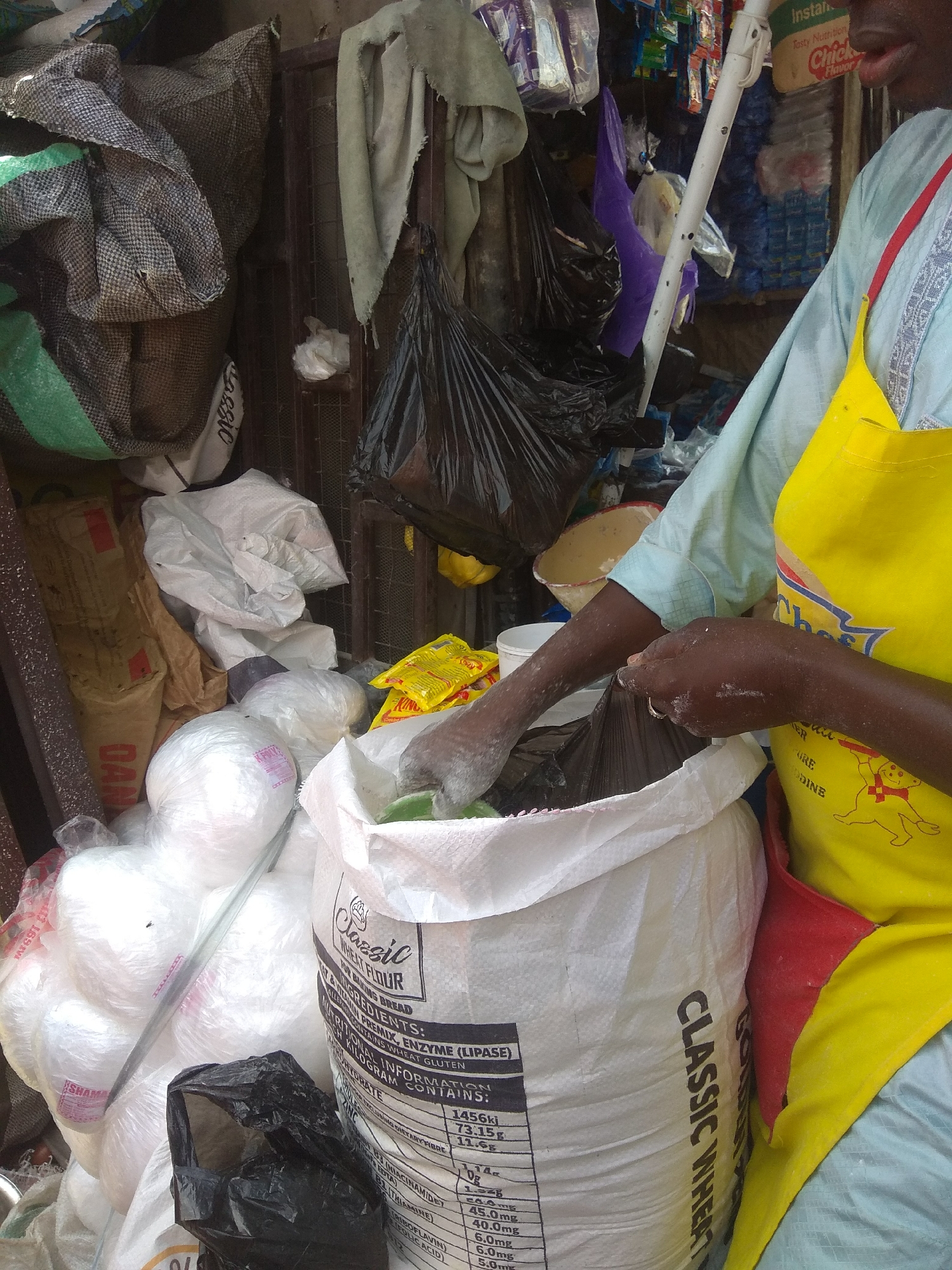 In a bustling marketplace setting in Jalingo, Nigeria, a person wearing a light blue garment and a vibrant yellow apron is actively engaged in handling a large white sack of wheat flour. The individual's hands, dusted with flour, are visible as they reach into the sack, suggesting they are scooping or measuring the flour. The surrounding environment is a typical market scene, characterized by an abundance of goods, many of which are also packaged in sacks and plastic bags.
Several other sacks, some appearing to be made of burlap and others of white plastic, are piled high around the person, creating a sense of a well-stocked stall. Among the goods, a black plastic bag is prominently displayed in the foreground, and various colorful packets and products are visible in the background, hinting at the diverse merchandise available. The lighting suggests it might be daytime, with no specific indications of weather conditions.
Visible text on the flour sack includes "CLASSIC WHEAT FLOUR FOR BAKING BREAD," along with a list of ingredients and nutritional information, including values for "ENERGY," "CARBOHYDRATE," "ZINC," and various vitamins such as "NIACINAMIDE" and "THIAMINE." The overall impression is one of a busy commercial exchange, with the person in the apron appearing focused on their task within this lively market.