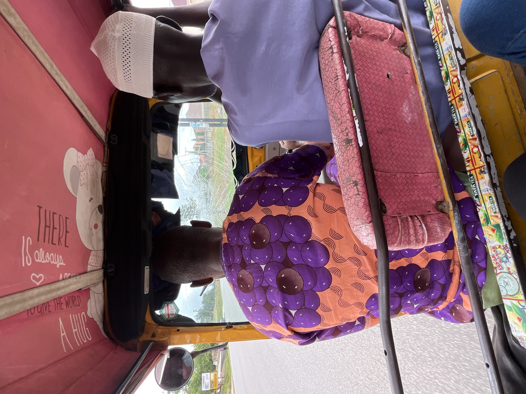 The view from inside an auto rickshaw (tuk-tuk) reveals two passengers, one with a white kufi cap and light purple shirt, and another wearing a vibrant orange and purple flower patterned shirt. The vehicle's interior shows pink decorative panels with cartoon motifs and the message "THERE is always A HUG TO GIVE THE WORLD." The rickshaw's frame is visible, along with seat cushions and a metal safety cage. Outside the tuk-tuk, cracks are visible in the windshield, and a street sign can be seen, suggesting a roadside setting with vegetation. TATA TEA branding on the vehicle suggests this scene occurs in Galadimawa, Nigeria.