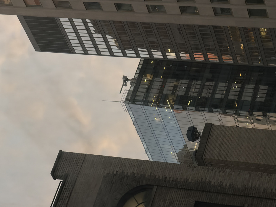 The image depicts a portion of an urban skyline in the City of New York, United States of America, against an overcast sky displaying faint grey and orange hues. Three distinct buildings are visible. On the far left, a brick-faced building, possibly an older structure, is partially framed, showing an arched window and a section of its upper structure with a rectangular, dark object atop a chimney-like feature. In the center, a modern skyscraper with a dark, reflective glass facade dominates, featuring a distinct curved or angled upper section. On its rooftop, a light-colored maintenance crane or window-washing apparatus and a slender antenna or lightning rod are present. Interior lights are visible through some of its windows, and its surface reflects parts of the sky and adjacent buildings. To the right, a taller, multi-story building with a light grey facade, likely concrete or stone, is partially visible. This building features a grid of numerous rectangular windows that also reflect portions of the environment, including warm, orange tones consistent with the sky. No individuals are depicted.