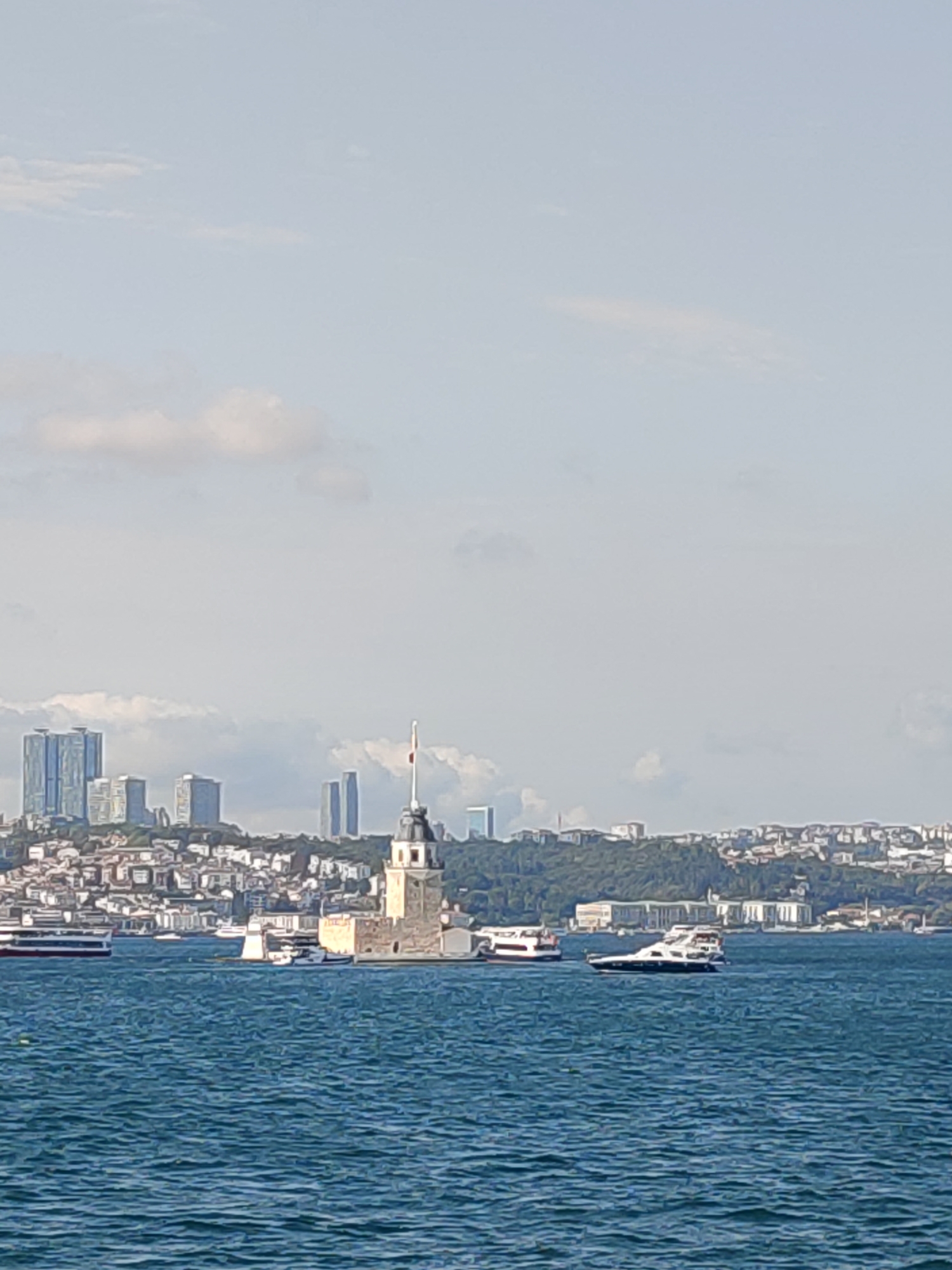 The image shows a distant view of the Maiden's Tower (Kız Kulesi) located in Üsküdar, Turkey. The tower is situated on a small islet in the Bosphorus strait. In the background, the skyline of Istanbul is visible, featuring several modern skyscrapers and other buildings. Several boats and ferries are present on the water, indicating maritime activity. The sky is partly cloudy, and the water is a deep blue. The overall scene depicts a sunny day with a clear view of the historical and modern landmarks of Istanbul.