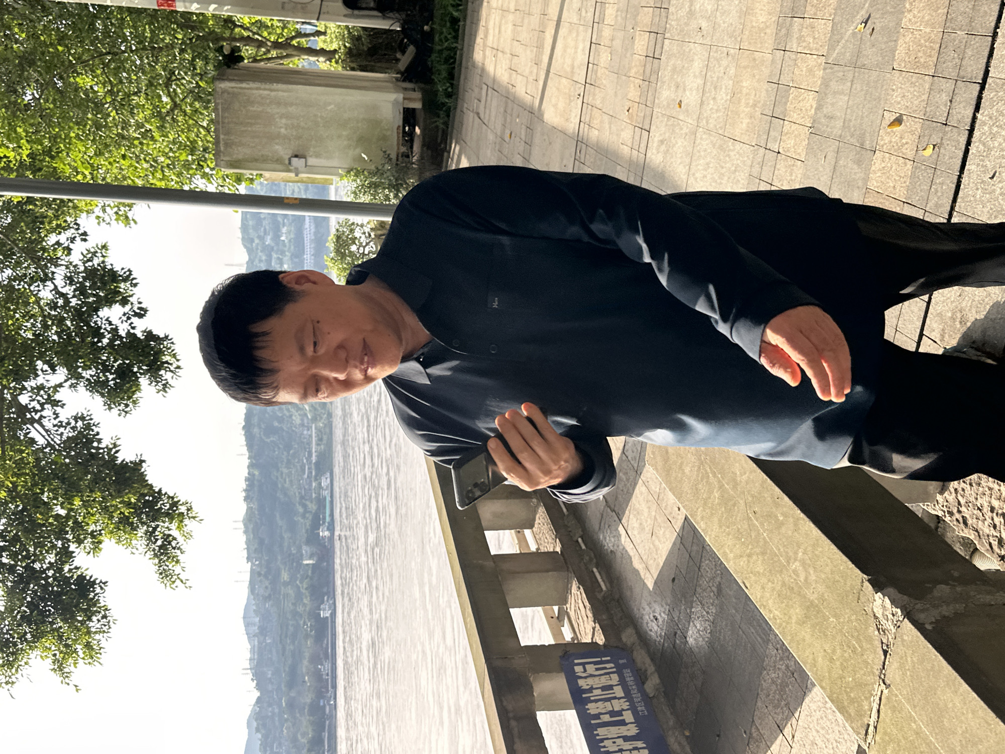A middle-aged Asian man is standing on a paved walkway, holding a smartphone. He's near a concrete barrier with a blue sign in Chinese. In the background, there's a wide river, possibly the Yangtze, with tree-covered hills on the far bank. The location is likely in or near Jiangjin, China, given the architecture and language on the sign. The man appears to be interacting with his phone, possibly taking a photo or looking at something on the screen. The scene is outdoors during the day with ample sunlight.
