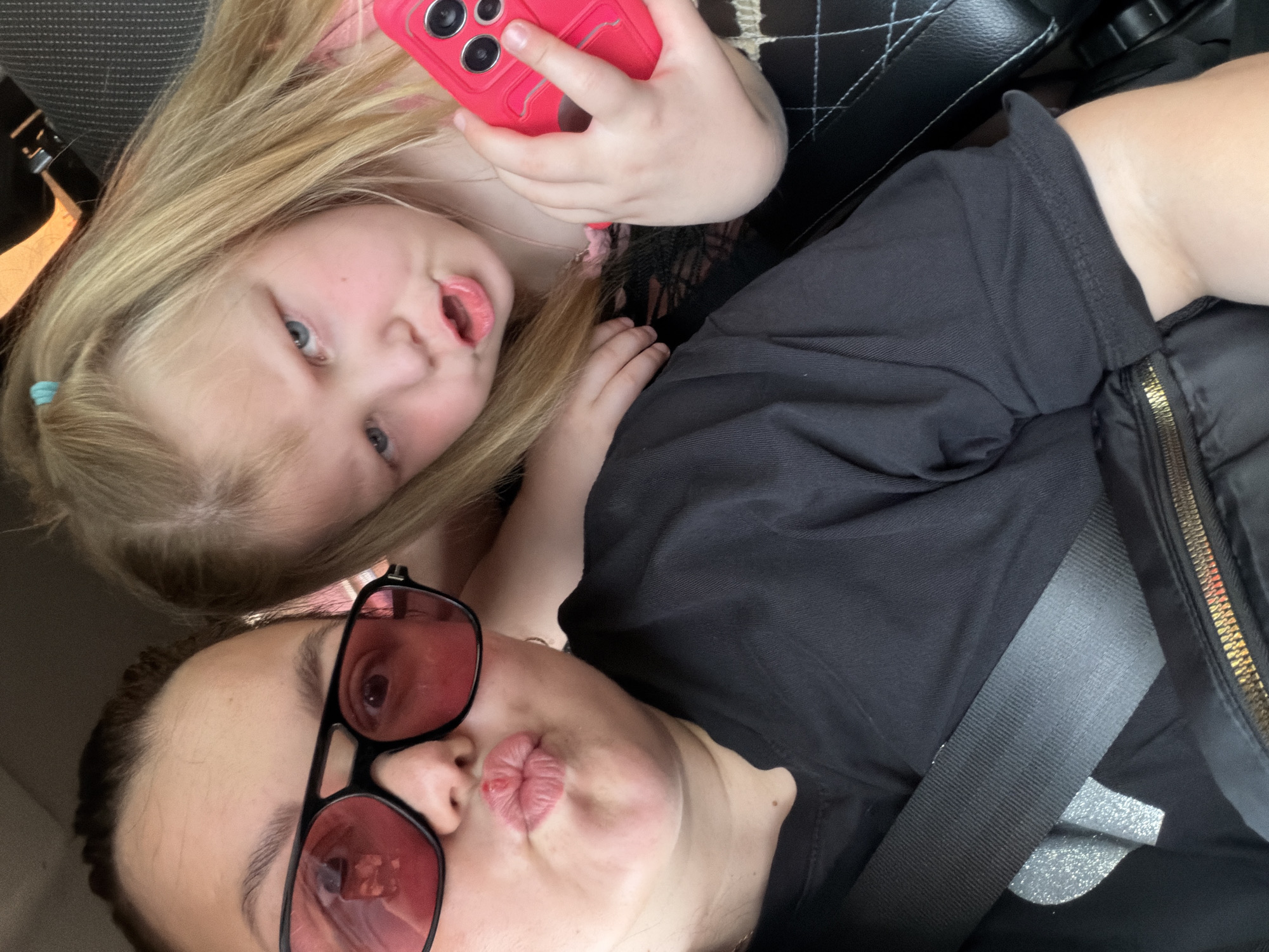 The media file shows a close-up, selfie-style photograph taken from a low angle. Two individuals are visible. In the foreground, a person wearing large, red-tinted sunglasses and a black t-shirt is puckering their lips, appearing to blow a kiss. Their facial expression suggests playfulness. Behind them, a child with blonde hair is also holding a pink smartphone and making a similar puckered expression. The child's hair is slightly messy, and they are wearing a pink top.
The setting appears to be inside a vehicle, possibly a car, suggested by the presence of a car seat headrest and a seatbelt in the background. The lighting is natural, indicating it could be daytime. The overall mood is casual and lighthearted, with both subjects engaged in a playful pose. The image is slightly tilted.
There is no visible text in the media file. The location context provided is Izhevsk, Russia, but there are no specific visual cues within the image to confirm this location.