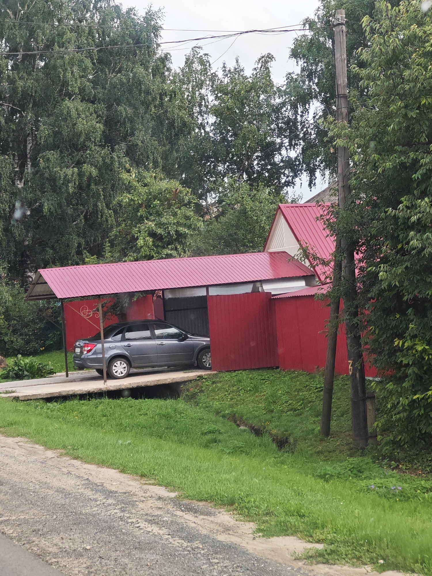 The image shows a grey sedan parked under a red-roofed carport. The carport is attached to a red building, and there is a red fence in the background. The scene is surrounded by trees and green grass. The ground in front of the carport is a gravel road. There are utility poles with wires visible in the background. The sky is overcast. The image appears to be taken in a residential area, possibly a dacha or country home, in Spassk, Russia.