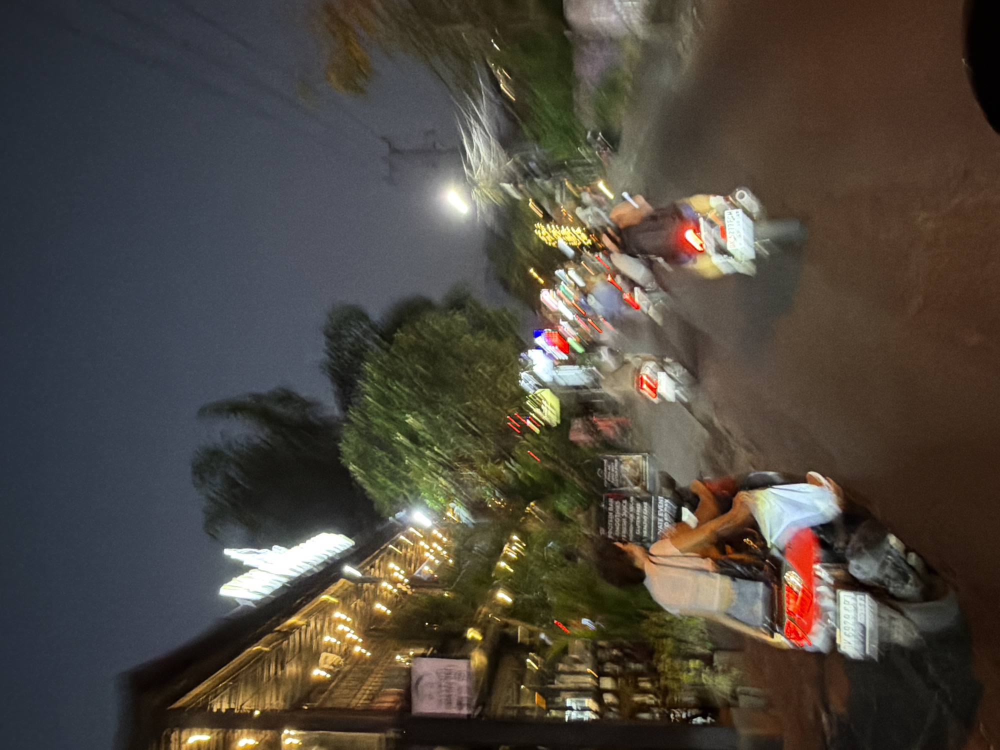This image presents a nocturnal street scene in North Kuta, Indonesia, characterized by significant motion blur, primarily affecting moving elements. On the left side of the frame, a multi-story building with a rustic, possibly wooden or bamboo, facade is brightly illuminated by numerous warm-toned string lights draped along its overhangs. A prominent, neon-like sign on the building's roofline is visible, though its text is blurred; the characters "WARUNG" appear partially discernible. Below, a legible sign lists offerings including "PROTEIN BAR," "SMOOTHIES," "FRESH JUICE," "HOMEMADE VEGAN," "GLUTEN FREE," "HEALTHY CAKE," and "PRIVATE EVENT." Green foliage and trees are visible adjacent to the building and lining the street.
The asphalt road is busy with several motorcycles. In the immediate foreground on the left, a person, likely female with short, dark hair and wearing a light top and pants, is seated as a passenger on a motorcycle. The motorcycle's red tail light is illuminated, and its license plate clearly displays "DK 6929 CPI." Further down the road, another motorcycle in the mid-ground has a visible license plate reading "DK 5277 PCW." Multiple other motorcycles and vehicles are represented by streaks of light and blurred forms, indicative of movement. A bright street light illuminates the mid-ground. The sky overhead is dark, consistent with a night environment.
