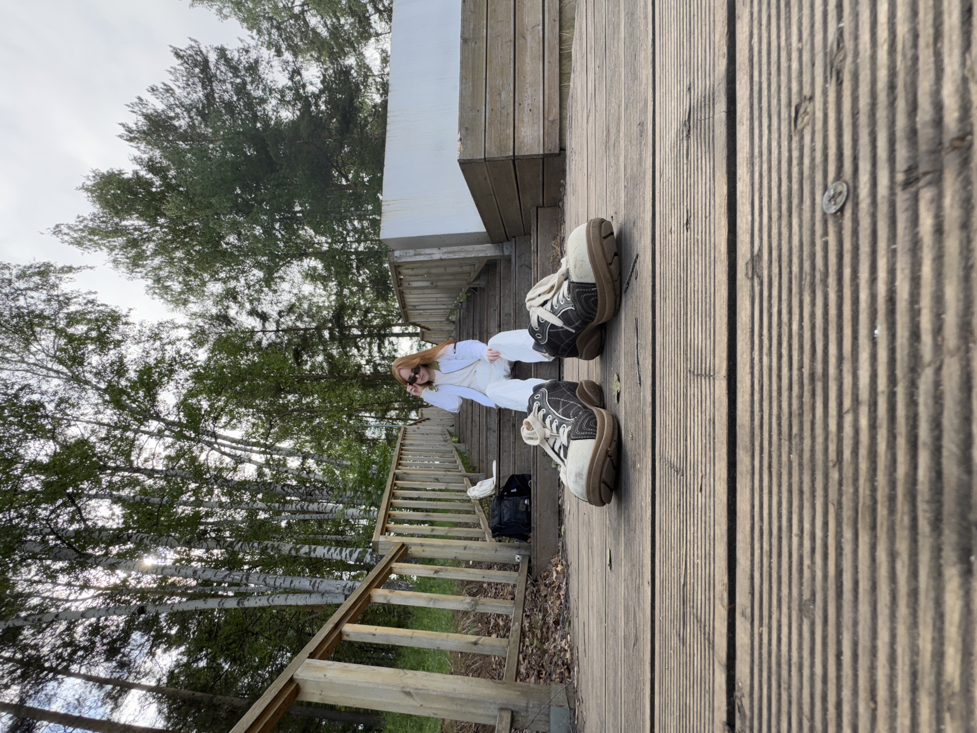 A medium, outdoor shot depicts a person in white attire seated on wooden steps, with an emphasis on the person's shoes, taken in what appears to be a wooded area with birch trees. The location is potentially in the city of Chaikovsky, Russia. The individual is wearing sunglasses. A black bag is placed on the steps behind them. The steps feature a wooden railing on the left side, leading down into the wooded area. A wooden surface is next to the steps. Two worn shoes with black and white detailing are placed on the surface. The background is filled with trees and sky.