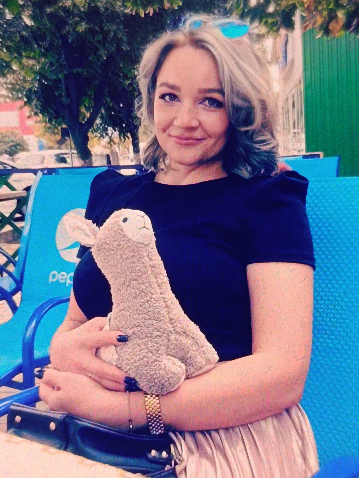 A woman with blonde hair, styled with waves and teal highlights, is holding a stuffed alpaca toy. She is wearing a dark blue top and a metallic silver skirt. She is seated on a bright blue chair with a Pepsi logo visible on its back. In the background, there is a large tree with green foliage and a glimpse of a green corrugated metal structure. The setting appears to be an outdoor seating area, possibly in a park or an establishment with outdoor seating in Bălți, Moldova.