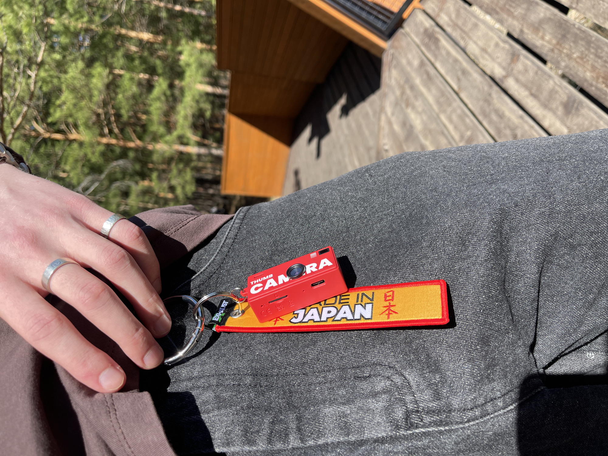 A medium-shot photograph depicts a person seated outdoors on a wooden surface, possibly a deck or patio, with their legs crossed. The focus is primarily on the person's left hand, which rests on their lap near the left side of the frame. The hand has two silver rings, one on the ring finger and one on the pinky finger. A red keychain, consisting of a miniature camera model and a yellow tag with red trim displaying "MADE IN JAPAN" in English and Japanese characters, lies on the person's left leg. The leg is clad in gray denim jeans. The person is wearing a brown shirt.
The background reveals glimpses of a building's wooden exterior, along with verdant trees. The environment suggests an outdoor setting in a natural or semi-urban area. The location details (Krasnoyarsk, Russia) are not evident in the image.