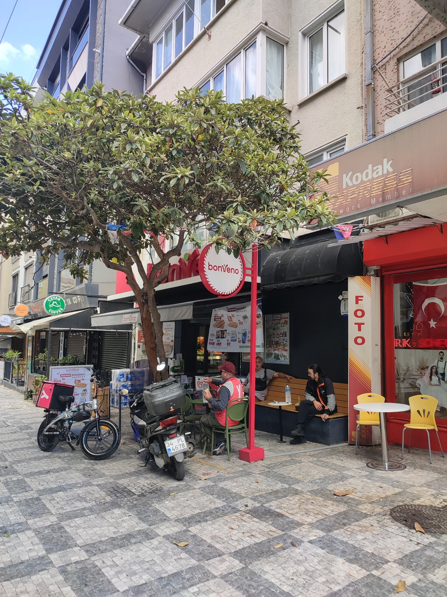 The image depicts a street scene in Kadıköy, Turkey. In the foreground, a gray and white checkered pavement leads to a row of businesses. A large, leafy tree dominates the center of the frame, with its branches extending over the sidewalk. To the left, a foldable electric bicycle with a red delivery box is parked. Next to it, a motorcycle with a large black storage box and a license plate reading "34 PL 4678" is parked.
Several people are present. Two men are sitting on a wooden bench outside a shop. One man, wearing a red vest and cap, is holding food. The other man, in a black and orange jacket, is seated next to him. Another person is visible through the shop window, possibly a customer.
Above the shops, building facades are visible. One building has a prominent sign that reads "Kodak EXPRESS Network Member." Another shop features a red awning and a sign with a circular logo and the text "bonVeno." A vertical sign with "FOTO" written in red letters on a yellow and orange background is also visible. The overall environment suggests a commercial area with a mix of food establishments and service providers. A Turkish flag is displayed inside one of the shops.