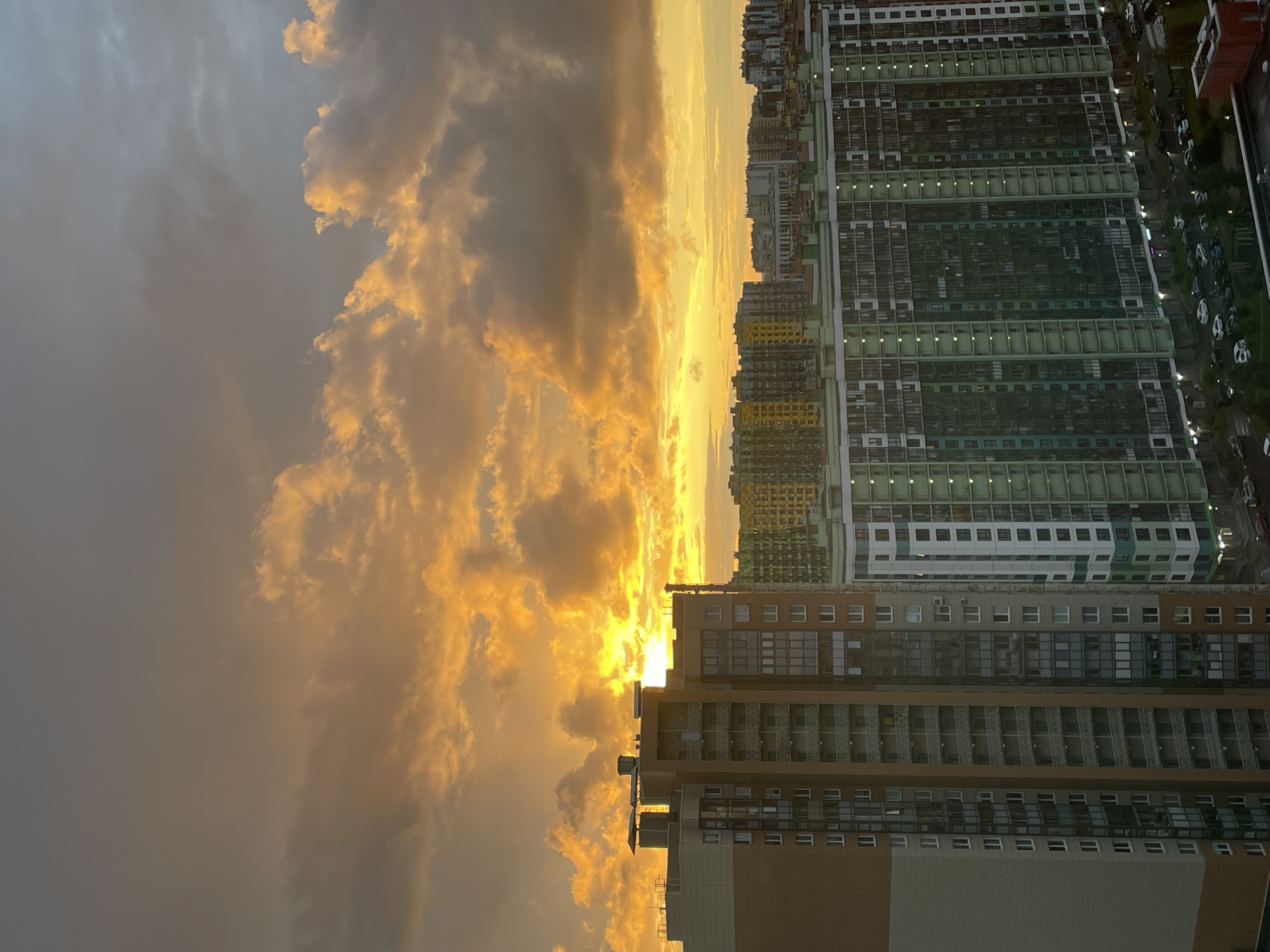 The image depicts a sunset over a residential area in Saint Petersburg, Russia. The sky is filled with dramatic, illuminated clouds, casting a warm, golden light. Several multi-story apartment buildings, characteristic of modern urban development in the city, dominate the foreground and midground. These buildings feature a variety of architectural styles and color palettes, with some displaying balconies and numerous windows. In the lower portion of the image, cars are visible parked on the streets and in lots around the buildings, indicating an active residential environment. The overall scene captures a moment of urban twilight.