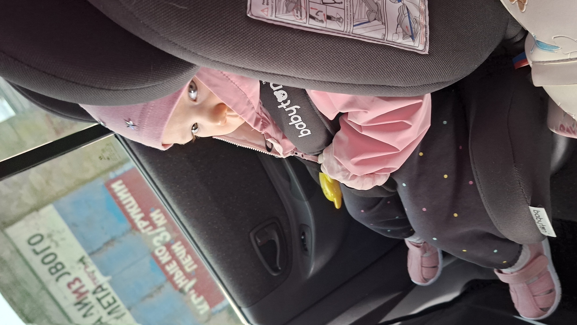 A young child, presumably a baby or toddler, is sitting in a car seat inside a vehicle. The child is wearing a pink hat and jacket, along with dark-colored pants adorned with polka dots. The car seat is gray and has the word "babyton" on the harness strap. The child is also wearing pink sandals. The interior of the car is visible, including the door handle and car seat instructions. Through the car window, there is an outdoor scene, and visible on the side of a building or structure are Russian words that could be identified as being in the city of Chaikovsky, Russia.