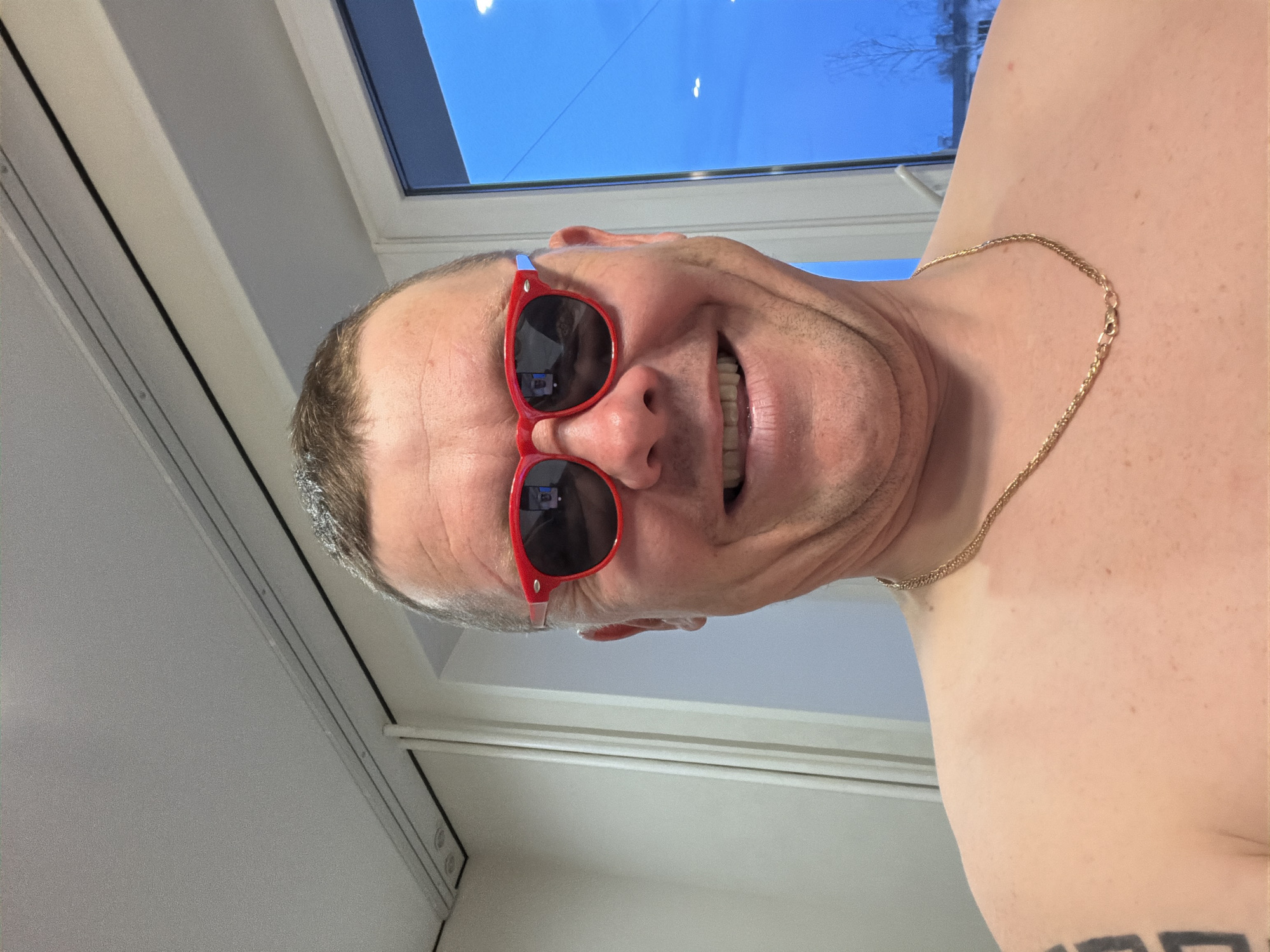 A rotated portrait-oriented shot captures a shirtless, middle-aged man with short, greying hair, smiling broadly and showing his teeth. He is wearing red-framed sunglasses, which reflect an indoor environment and possibly the camera taking the picture. A gold chain is visible around his neck, and a black-ink tattoo is partially visible on his right upper arm.
The man's head is positioned on the left side of the frame, tilted downwards due to the image orientation. He looks directly towards the viewer with an expression of happiness or amusement. In the background, a window reveals a clear blue sky and a few bare tree branches, indicating bright daytime conditions. The indoor setting appears to be a room with light-colored walls and window frames. The image depicts a personal moment, likely a selfie, taken indoors in Chaikovsky, Russia. No visible text is present in the image.