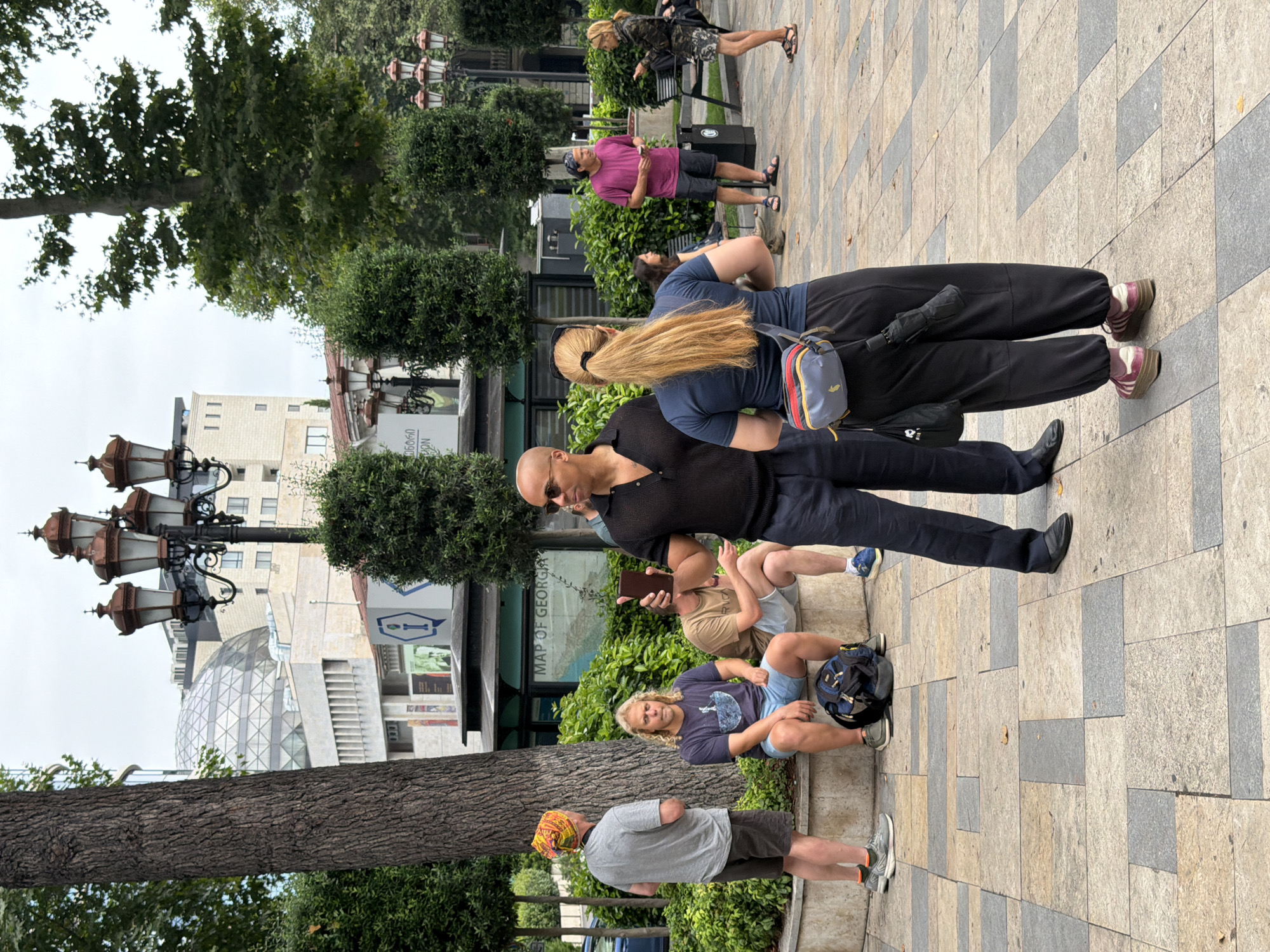 A group of people are gathered in an outdoor public space in Tbilisi, Georgia, possibly Rustaveli Avenue, based on the architectural style and the presence of what appears to be a "Map of Georgia" kiosk. A bald man wearing sunglasses and a black polo shirt is in the foreground, holding a dark-colored object, possibly a phone, in his right hand. To his left, a woman with long blonde hair in a ponytail wears a dark blue t-shirt, black pants, and a grey, red, and blue crossbody bag. She appears to be looking towards the bald man.
Further back, to the left of the bald man, three men are seated on a low wall. One man wears a yellow and orange patterned head covering, a grey t-shirt, and dark shorts. Next to him, a man with blond hair wears a dark blue t-shirt and light blue shorts. A third man, partially obscured, wears a tan t-shirt and light shorts. A blue backpack rests on the ground near the seated men.
In the background, a prominent geodesic dome structure is visible atop a modern building, flanked by more traditional buildings with ornate street lamps. A kiosk with "MAP OF GEORGIA" signage is situated behind the seated individuals. Other individuals are visible further in the background, including a person in a magenta shirt and dark shorts, and a woman in a camouflage jacket. The ground is paved with large rectangular stones in a geometric pattern. Green bushes and trees provide a backdrop to the scene.