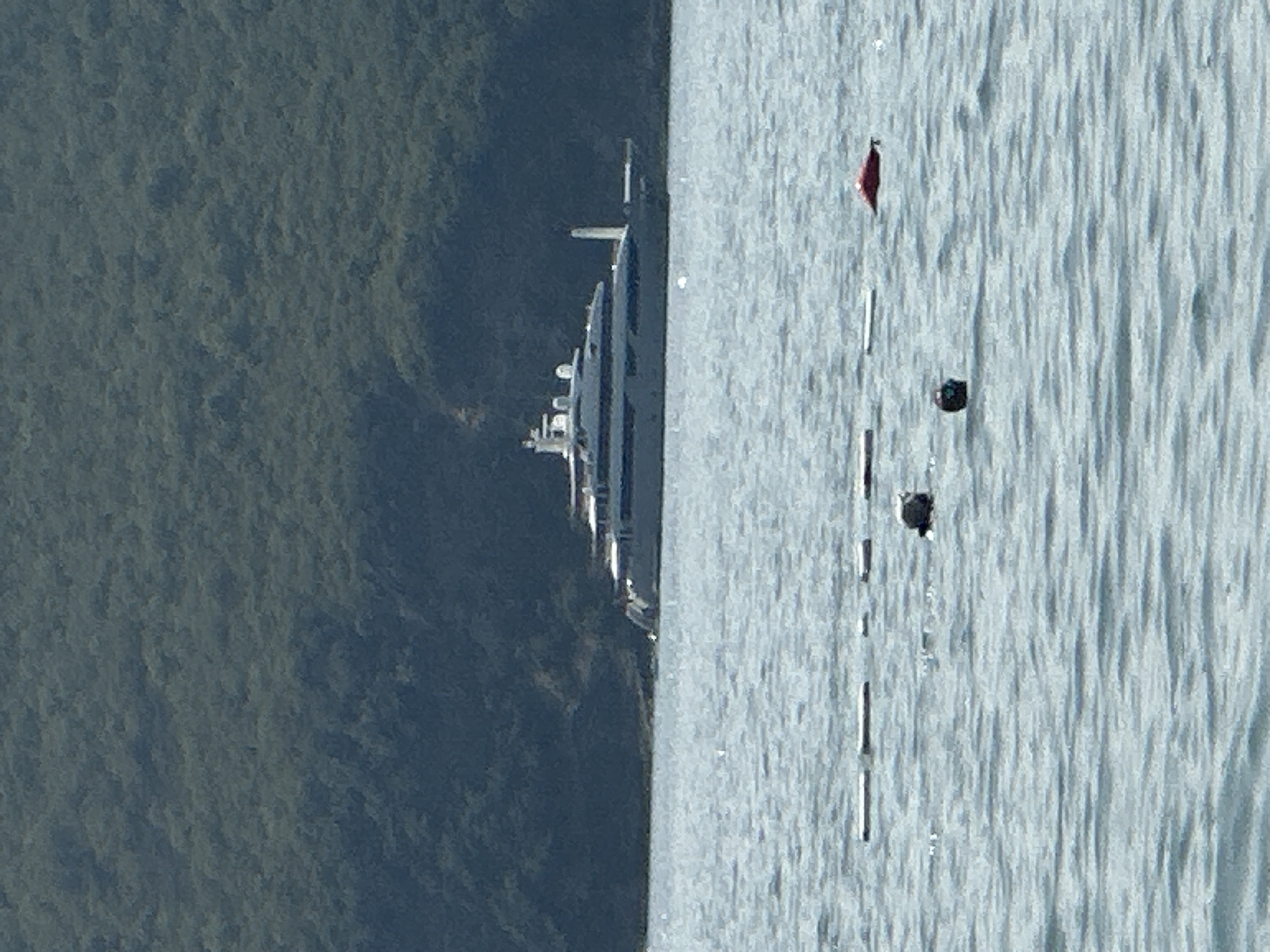 A large, white luxury yacht is anchored in the Bay of Kotor, near the town of Boreti, Montenegro. The yacht has multiple decks and several prominent structures on its upper levels, including what appears to be a helipad at the stern. The yacht is positioned in calm, blue-green water, with a lush, tree-covered hillside in the background. In the foreground, several buoys are visible on the surface of the water, marking a designated area. The sky is clear and bright, suggesting a sunny day.