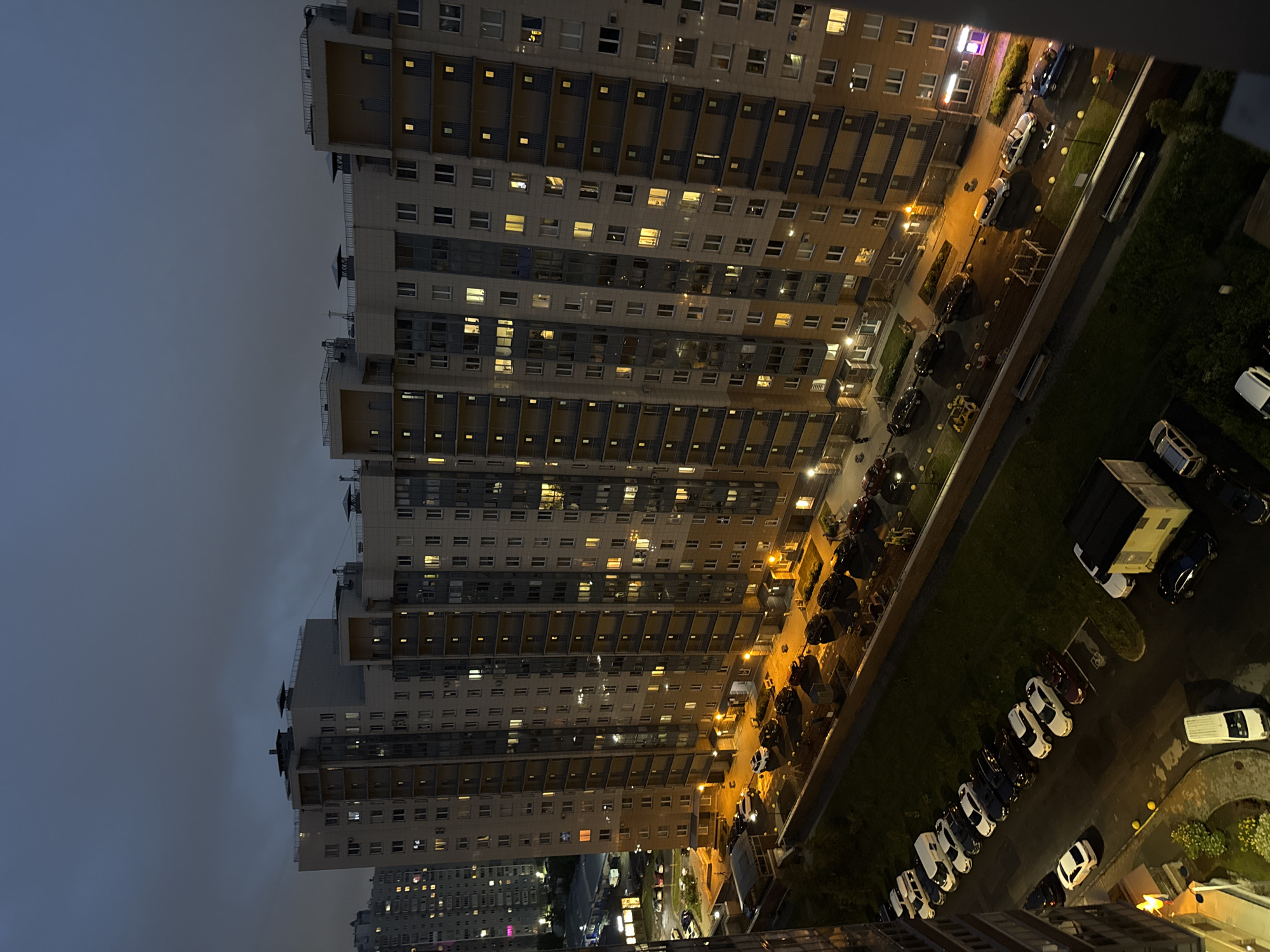 The image depicts a nighttime aerial view of a residential building in an urban environment. The building is a tall, multi-story structure with numerous windows, many of which are illuminated, suggesting habitation. In front of the building, a parking lot is visible, containing a variety of cars. A grassy area separates the parking lot from a sidewalk or pathway. Beyond the immediate area, other buildings and the sky are visible. The overall scene conveys a sense of a typical urban residential setting at night, with artificial lighting illuminating the scene against the dark sky.