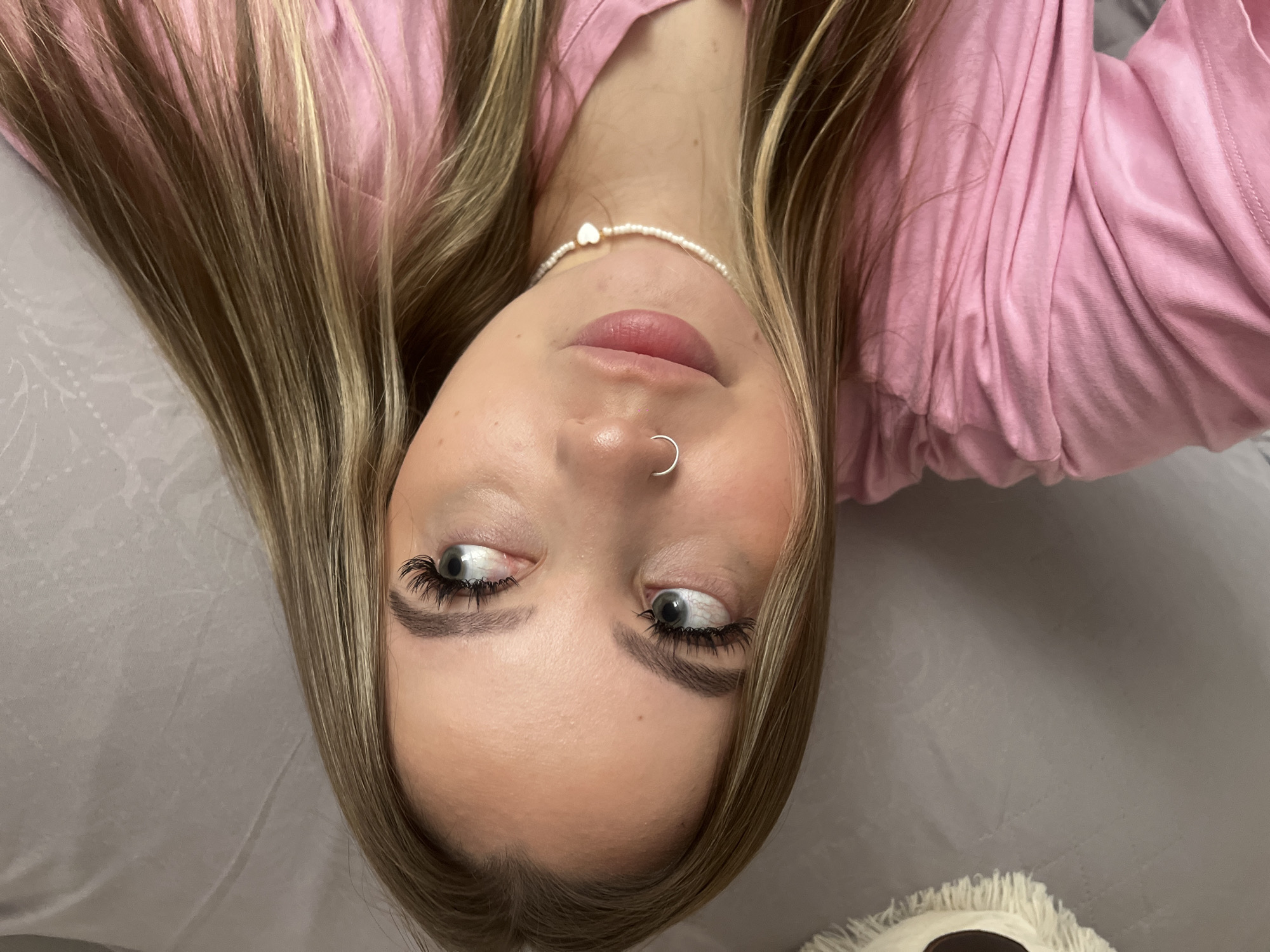 A young woman with blonde hair and a nose ring is lying on a grey bed. She is wearing a pink shirt and a pearl necklace with a heart pendant. Her eyes are looking towards the upper right of the image. Part of a plush toy is visible in the lower right corner. The location is not discernible from the image.