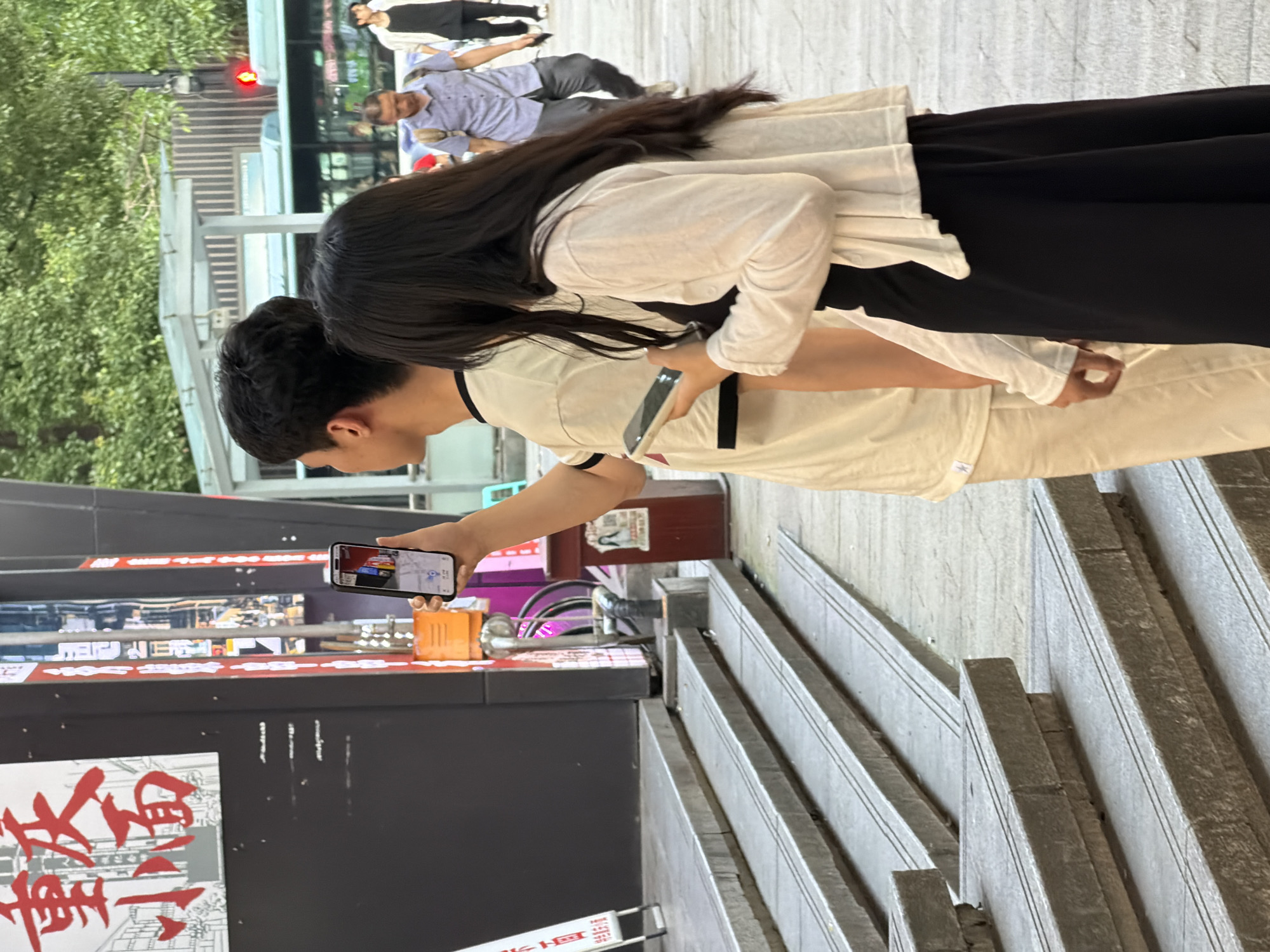 In Yuzhong District, China, on a street with a bus in the background and lush green trees overhead, a young man and woman stand side-by-side. The man, wearing a white t-shirt, holds a smartphone in his right hand, seemingly capturing a photo or video. The woman, dressed in a black skirt and a white cardigan, looks down at her own smartphone held in her left hand. The foreground features a set of concrete stairs leading up to a building with a dark facade and red and white signage. The scene depicts a casual moment on a city street, with both individuals engaged with their mobile devices.