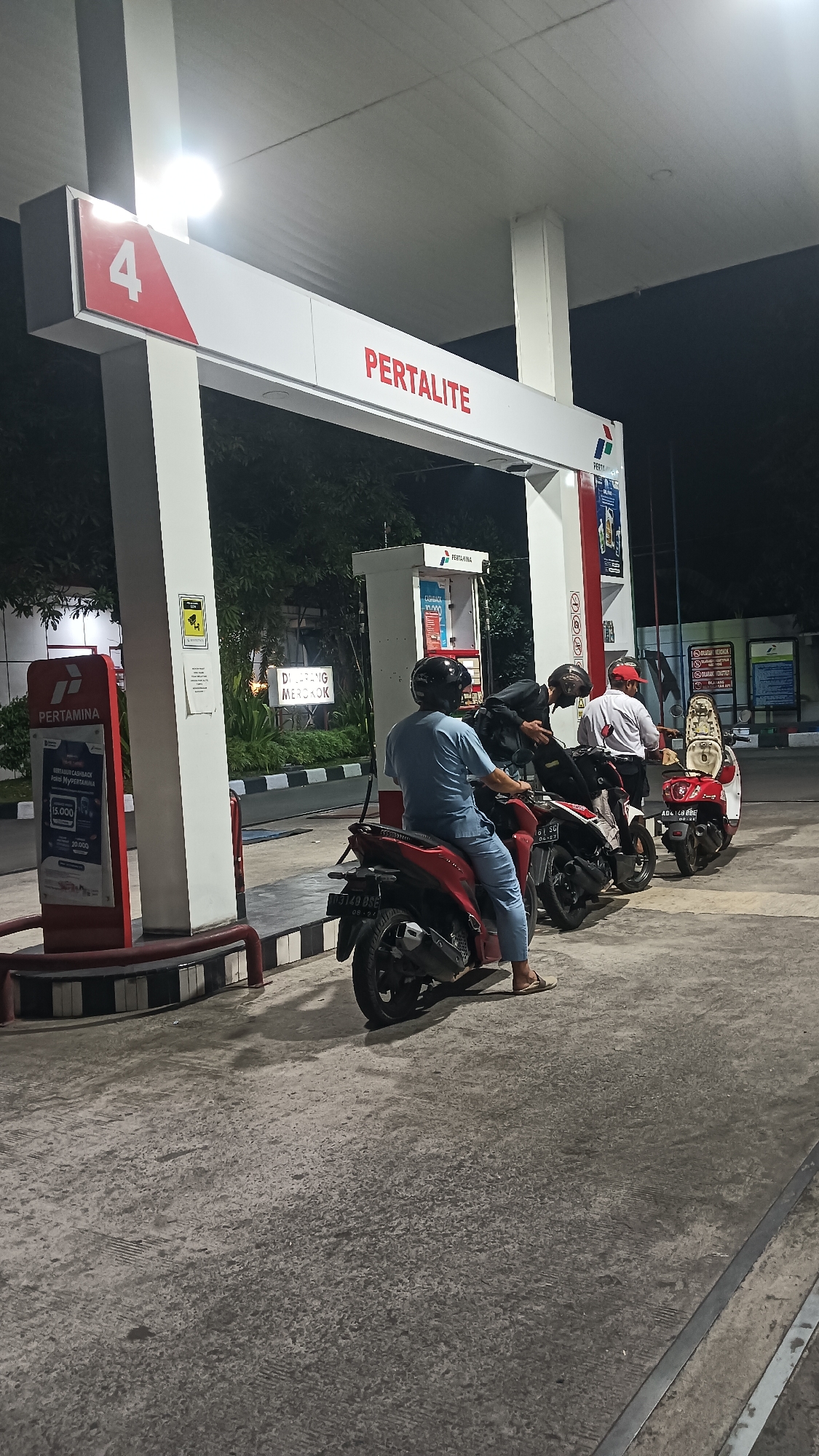 At a Pertamina gas station in Sambungmacan, Indonesia, four individuals on motorcycles are lined up at pump number four, which is labeled for "PERTALITE." The scene is dimly lit, suggesting it is nighttime. The ground is concrete, and the gas station canopy is supported by white pillars. One person in the foreground is wearing light blue medical scrubs and a black helmet. Another person behind them is wearing a black jacket and a helmet. A third person, wearing a red cap and white shirt, is standing near a motorcycle. The fourth person is obscured by the motorcycles. There are signs visible, including "PERTAMINA" logos and other promotional or informational text. A sign in the background reads "DILARANG MEROKOK," meaning "NO SMOKING." The environment appears to be an outdoor public space with some trees visible in the background.