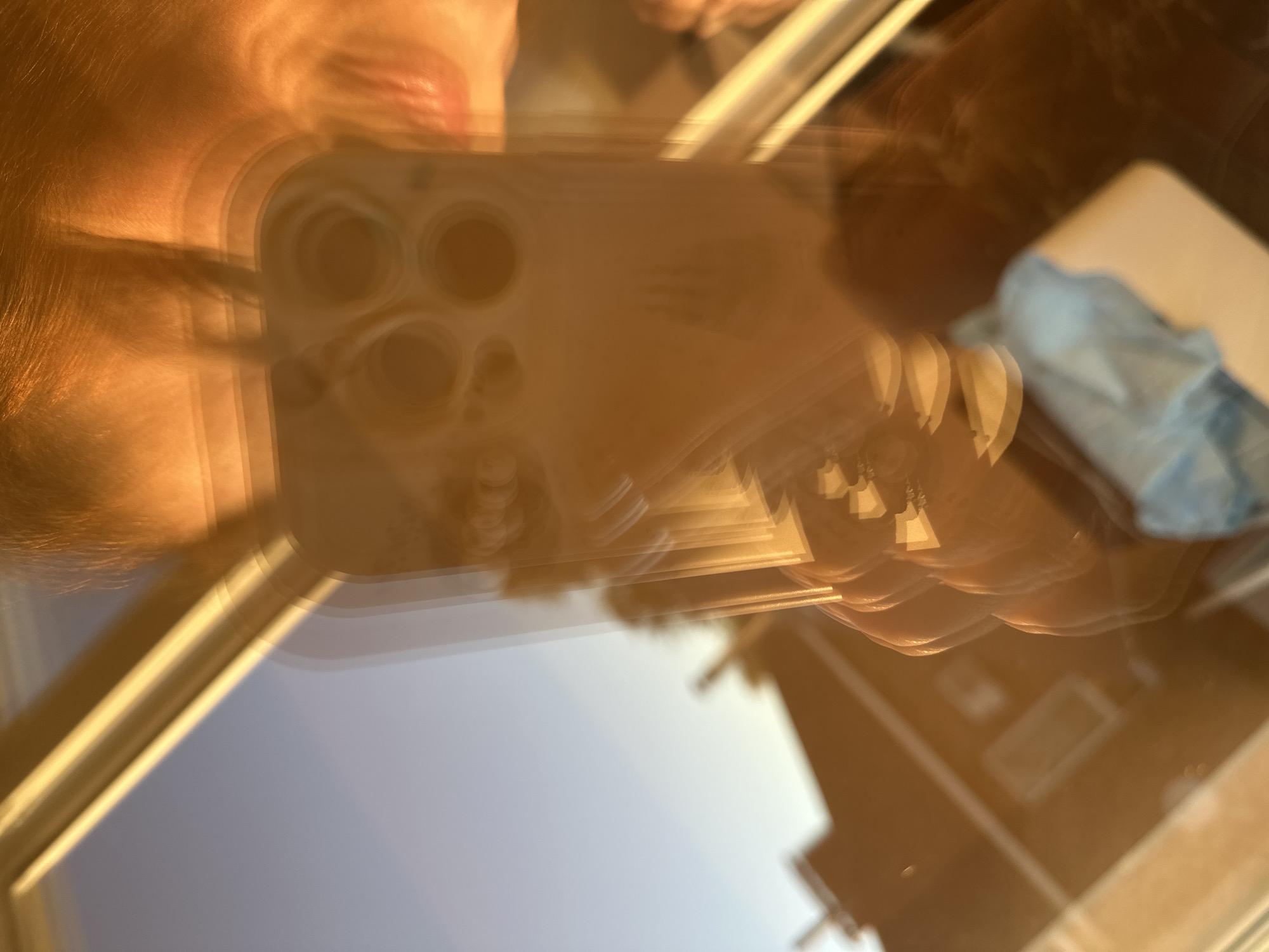 This is a close-up, motion-blurred photograph taken from a low angle, likely reflecting off a shiny surface such as glass or a mirror. The primary subject appears to be a person's hand holding a smartphone with a distinctive triple-lens camera array, possibly an iPhone. The hand and phone are seen in multiple overlapping exposures, creating a dynamic, almost ghostly effect. The warm, golden light suggests it is either sunrise or sunset, casting long shadows and highlighting the textures of the surface. In the background, hints of a room can be seen, including what looks like a light blue cloth or item on a surface and the blurred outlines of furniture or architectural elements. The overall impression is one of movement and transient light, capturing a fleeting moment. The location context of Nizhny Novgorod, Russia, is not visually discernible from the image itself. There is no visible text.