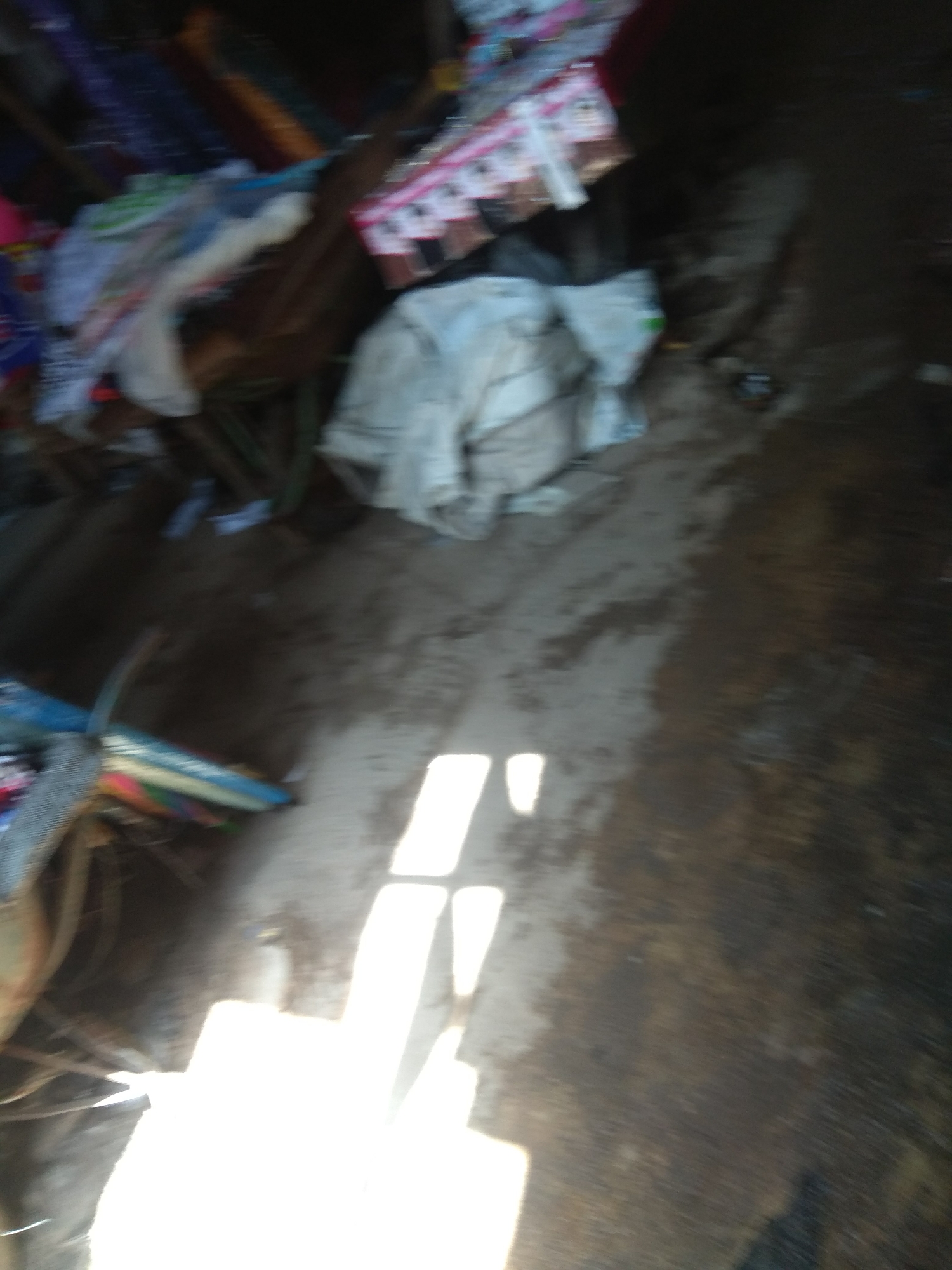 The media file shows a blurred, close-up, ground-level view of a messy, outdoor setting, possibly a market stall or a cluttered pathway. The scene is dimly lit, with strong, directional light creating bright white rectangular shapes on the ground, suggesting sunlight filtering through an overhead structure. The ground appears to be dirt or mud, with some areas looking wet.
Scattered across the ground and in piles are various objects. Some appear to be fabrics or textiles, with one particularly noticeable bundle of white cloth. There are also glimpses of what could be packaged goods, possibly in colorful wrappers or boxes, and some debris. The overall impression is one of disarray and perhaps neglect.
There are no clear indications of people or specific activities, beyond the implication of a place where goods are stored or displayed. The location context provided is Jalingo, Nigeria, which might suggest a tropical or subtropical environment, but the image itself doesn't offer strong visual cues to confirm this. The time of day is likely daytime due to the sunlight, but the quality of light suggests it could be early morning or late afternoon. No text is discernible in the image due to the blurriness. The mood conveyed is somewhat somber and disorganized.