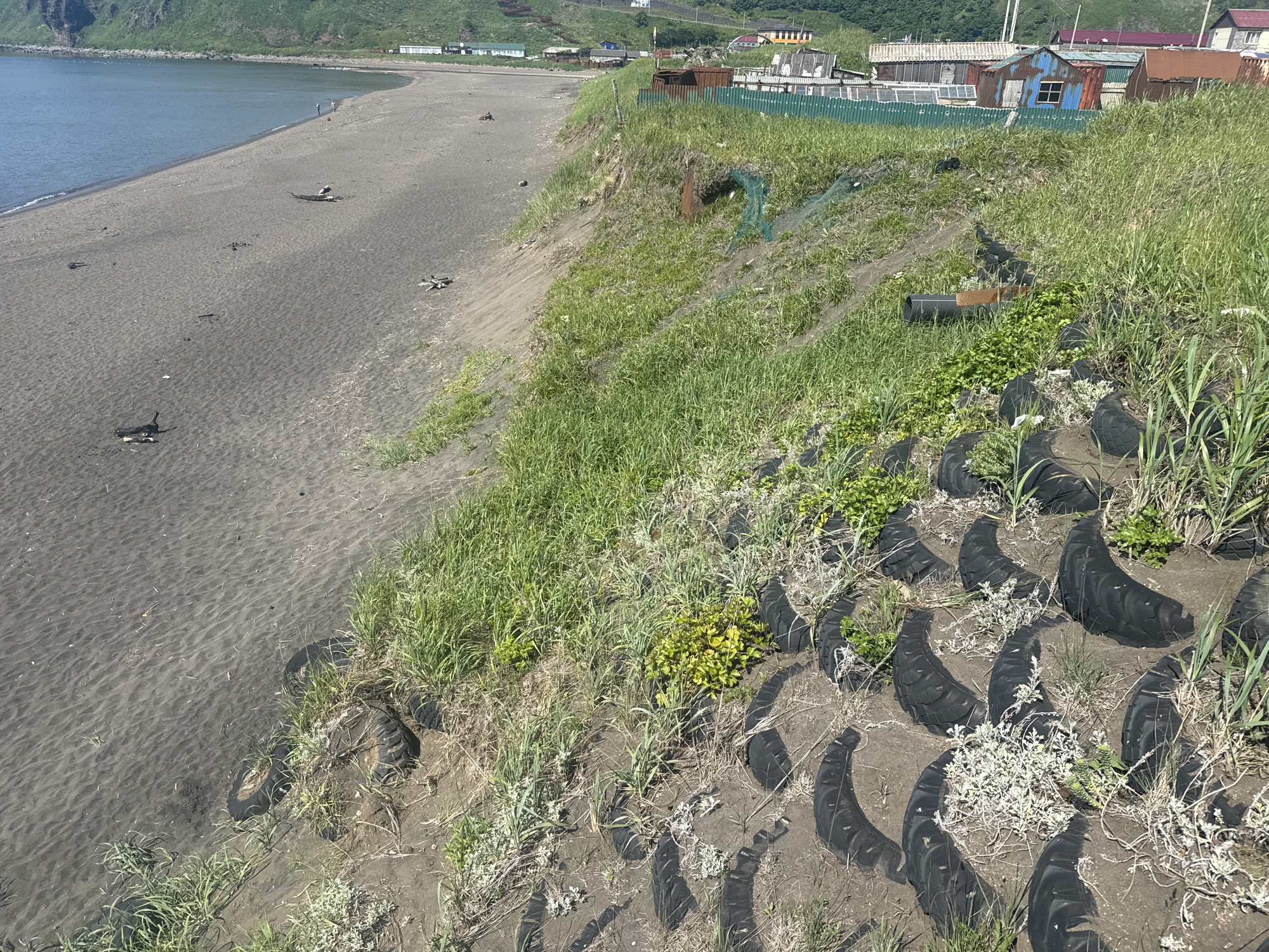 The image shows a black sand beach along the coast, with a grassy slope rising from the beach. Several old tires are placed along the slope, some partially buried in the soil and overgrown with grass. On the top of the slope, a few rustic buildings with corrugated metal roofs are visible, suggesting a small settlement. The sea is calm and blue. The location is consistent with the appearance of the outskirts of Kuril'sk, Russia, a city known for its volcanic beaches and coastal landscapes.