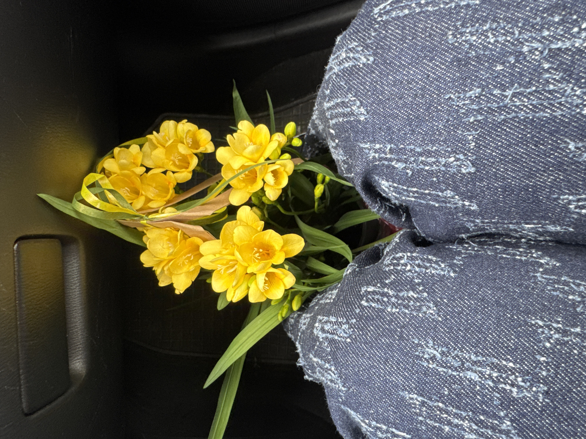 The image shows a bunch of yellow freesia flowers with green leaves and a decorative yellow ribbon. The flowers are placed in what appears to be the interior of a car, possibly a side compartment or dashboard area. Next to the flowers, there is a denim item, likely jeans, that is folded or draped. The location is not discernible. The image doesn't have enough information to determine if the scene occurs in Rudnik, Bulgaria.