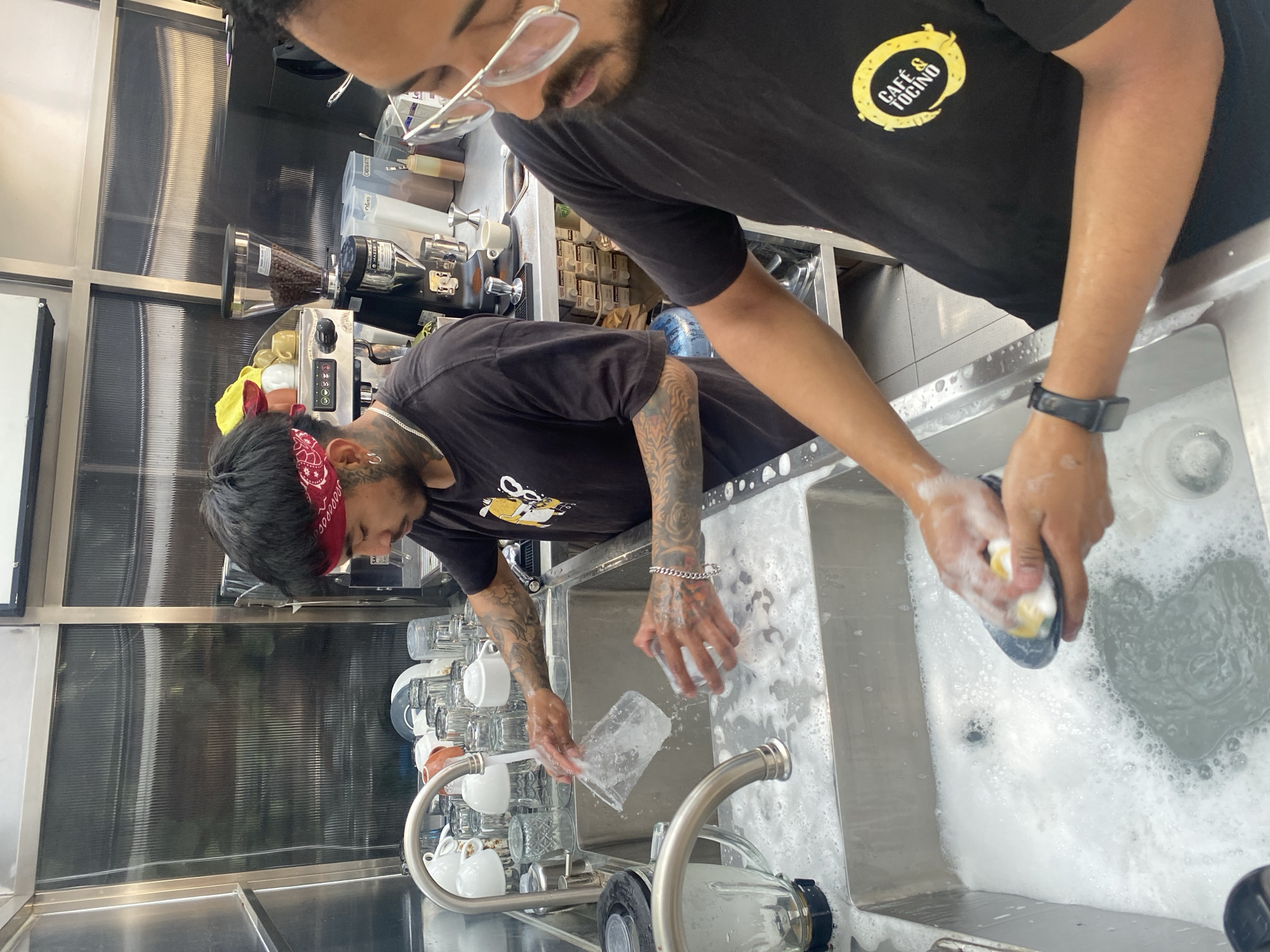 Two individuals are captured in a commercial setting, possibly a café in Puebla, Mexico. One man, adorned with a red bandana, tattoos on his arms, and a silver bracelet, is washing a clear cup in a stainless-steel sink. Another individual, wearing glasses and a black t-shirt featuring a logo that reads "Café & Tocino," is washing something with a sponge in the same sink. The sink is filled with soapy water. Behind them, a series of clean cups and an espresso machine are visible. The environment appears to be a bustling commercial kitchen.