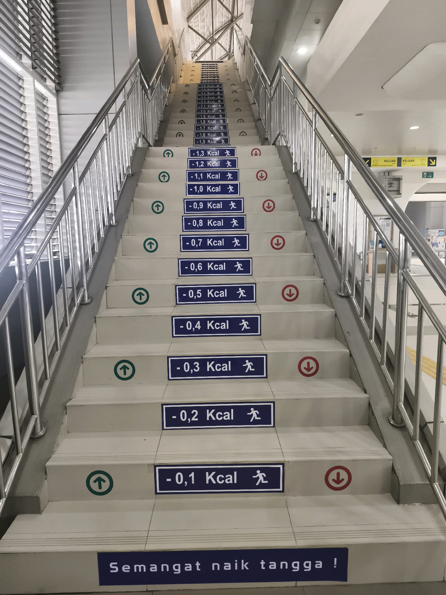 The image shows a set of concrete stairs with metal handrails in Palembang, Indonesia. Each step is marked with blue and white signs indicating calorie expenditure or the number of calories burned by taking that step. The signs show a number followed by "Kcal" and a small pictogram of a person walking or running. Some steps also have directional arrows, with green arrows pointing up and red arrows pointing down, indicating the direction of travel for calorie burning. At the bottom of the stairs, a blue sign reads "Semangat naik tangga!" which translates to "Keep climbing the stairs!" The environment appears to be a public building, possibly a train station or a mall, given the modern architecture and signage.