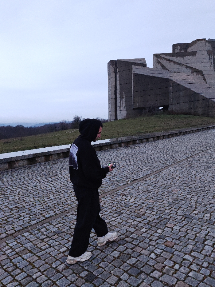 An individual is walking on a cobblestone pathway at the Monument to 1300 Years of Bulgaria in Shumen, Bulgaria, under an overcast grey sky. The person, viewed from the right side, is wearing a black hooded sweatshirt with a white and black graphic print on the back, black trousers, and off-white chunky sneakers. They are holding a smartphone horizontally with both hands at waist level, directing their gaze towards its illuminated screen. The pathway consists of dark grey, irregularly shaped cobblestones and slopes upwards. To the left of the path, a low grey concrete barrier separates it from a grassy embankment with sparse, leafless vegetation. In the background, the large, angular, brutalist concrete structures of the Monument rise from a grassy base, displaying weathering and dark streaks. Distant hills covered with trees are visible on the horizon to the left.