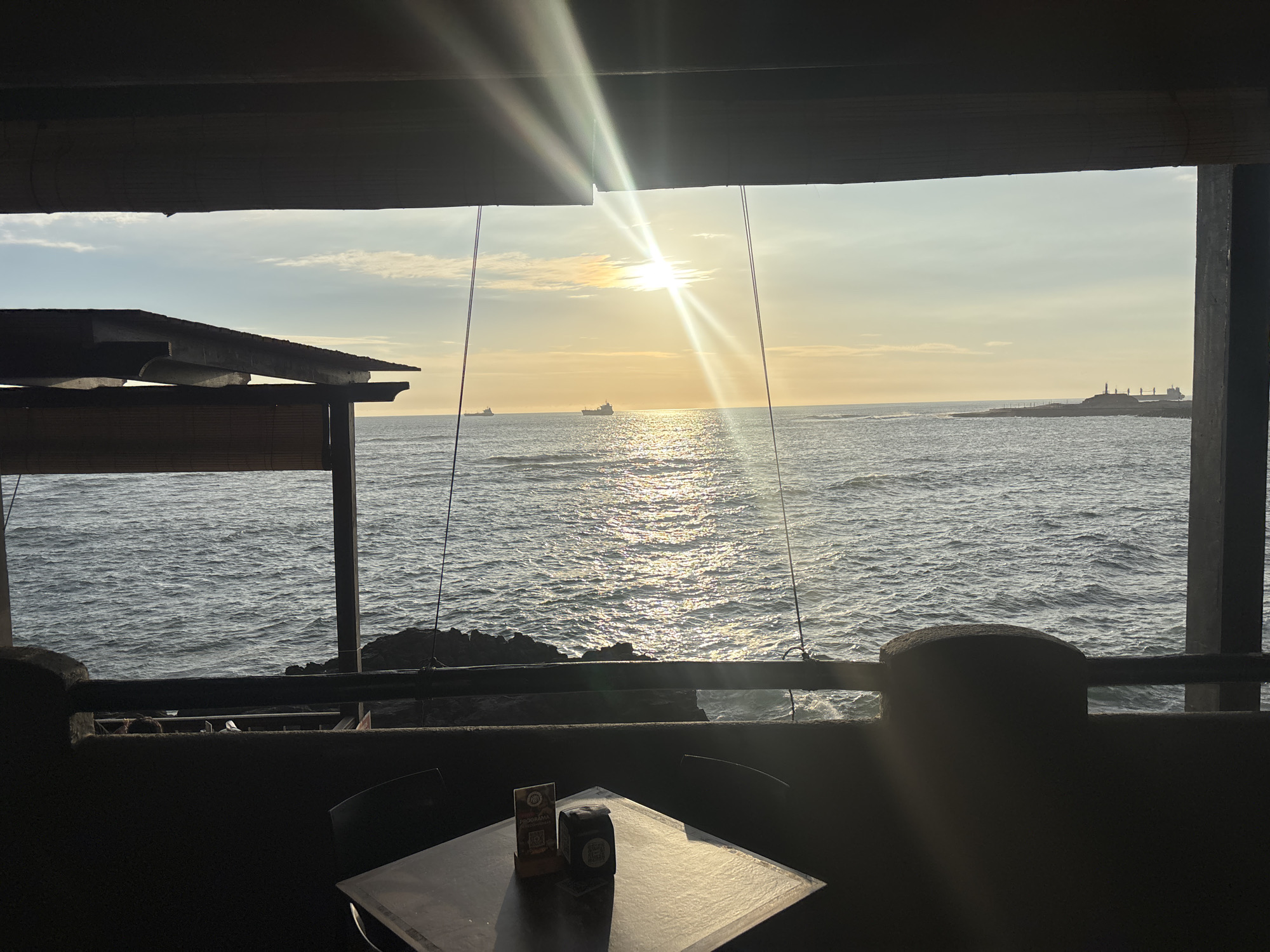 The image shows a view of the ocean through a partially enclosed space, likely a restaurant or patio. The sun is setting over the water, creating a bright glare. The ocean has moderate waves, reflecting the sunlight. Three ships are visible in the distance.
In the foreground, there's a table with a beverage container and a small menu or advertisement on it. There are two chairs around the table, indicating a space for dining. The structure is dark and appears to be constructed of wood and stone.
Based on the geographic location of Arica, Chile, and the presence of ships in the water, it can be assumed that this view overlooks the Pacific Ocean. The rocky shoreline visible in the middle ground suggests a coastal environment.
