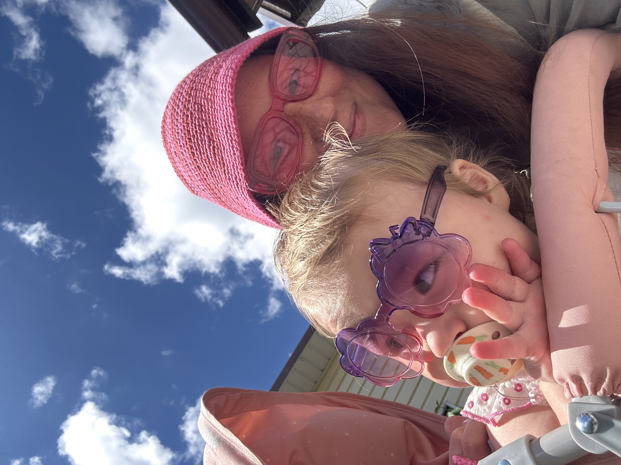 An adult woman and a young child are captured in a bright, upward-angled close-up shot, likely outdoors in Nizhny Novgorod, Russia. The woman, positioned at the top of the frame, has long, reddish-brown hair and wears a pink knitted hat and distinct red-framed sunglasses, which reflect the sky. Her expression is partially obscured by the glasses, but she appears relaxed.
Below her, a young child, possibly in a stroller or carrier, wears unique purple-framed sunglasses shaped like flowers. The child has light-colored hair and is holding a white pacifier decorated with orange carrot motifs in their hand and mouth. A portion of the child's pink clothing and the edge of a gray and pink stroller or carrier are visible.
The background is dominated by a clear, vibrant blue sky with scattered fluffy white clouds, indicating a sunny day with good weather. The edge of a building or structure, possibly a roofline or awning, is visible against the sky. The overall scene suggests a casual and cheerful outdoor moment. There is no visible text in the image.