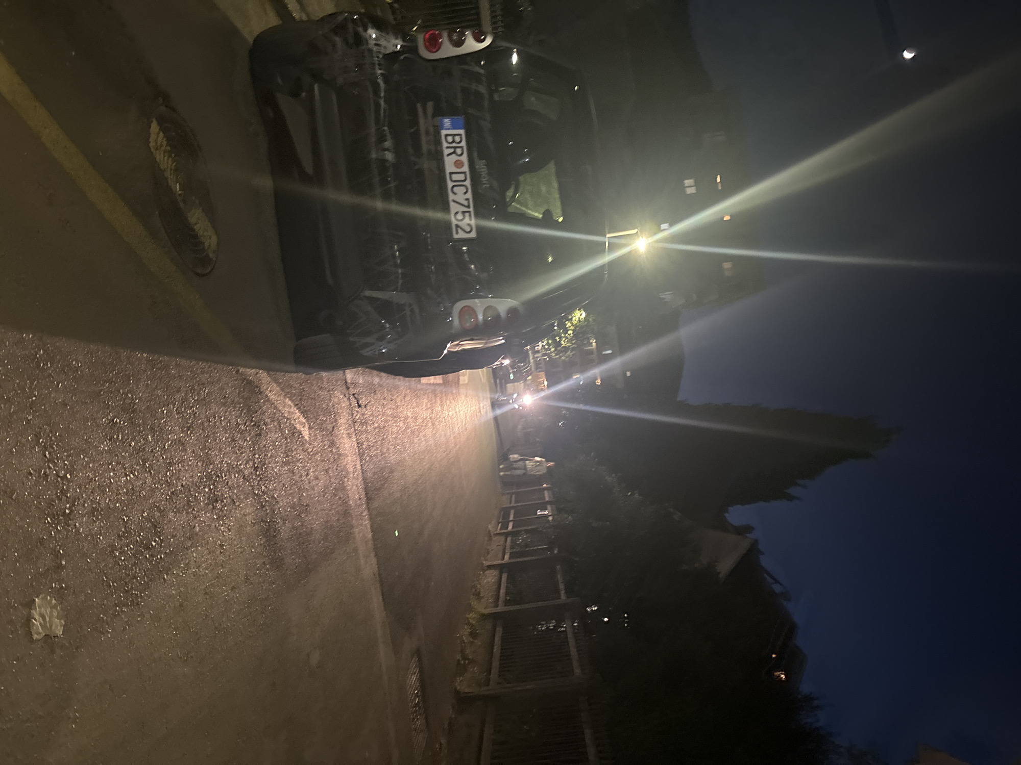 The image depicts a nighttime scene on a paved street in Budva, Montenegro. The foreground is dominated by the rear of a black Smart car, its license plate reading "BR DC752". The car appears to be parked or stopped, with its brake lights illuminated. The street surface is wet, reflecting the light from street lamps and the car's headlights, suggesting recent rain or humidity.
To the right of the car, a sidewalk runs alongside a wooden fence. Beyond the fence, trees and residential buildings are visible in the dim light. A single person, appearing to be a pedestrian, walks along the sidewalk in the distance, heading away from the viewer. Further down the street, another vehicle's headlights are visible.
In the upper left quadrant of the image, a sliver of the moon is visible in the dark sky, alongside bright streaks of light, likely lens flare from streetlights. The overall atmosphere is urban and nocturnal, with artificial lighting playing a significant role in illuminating the scene.