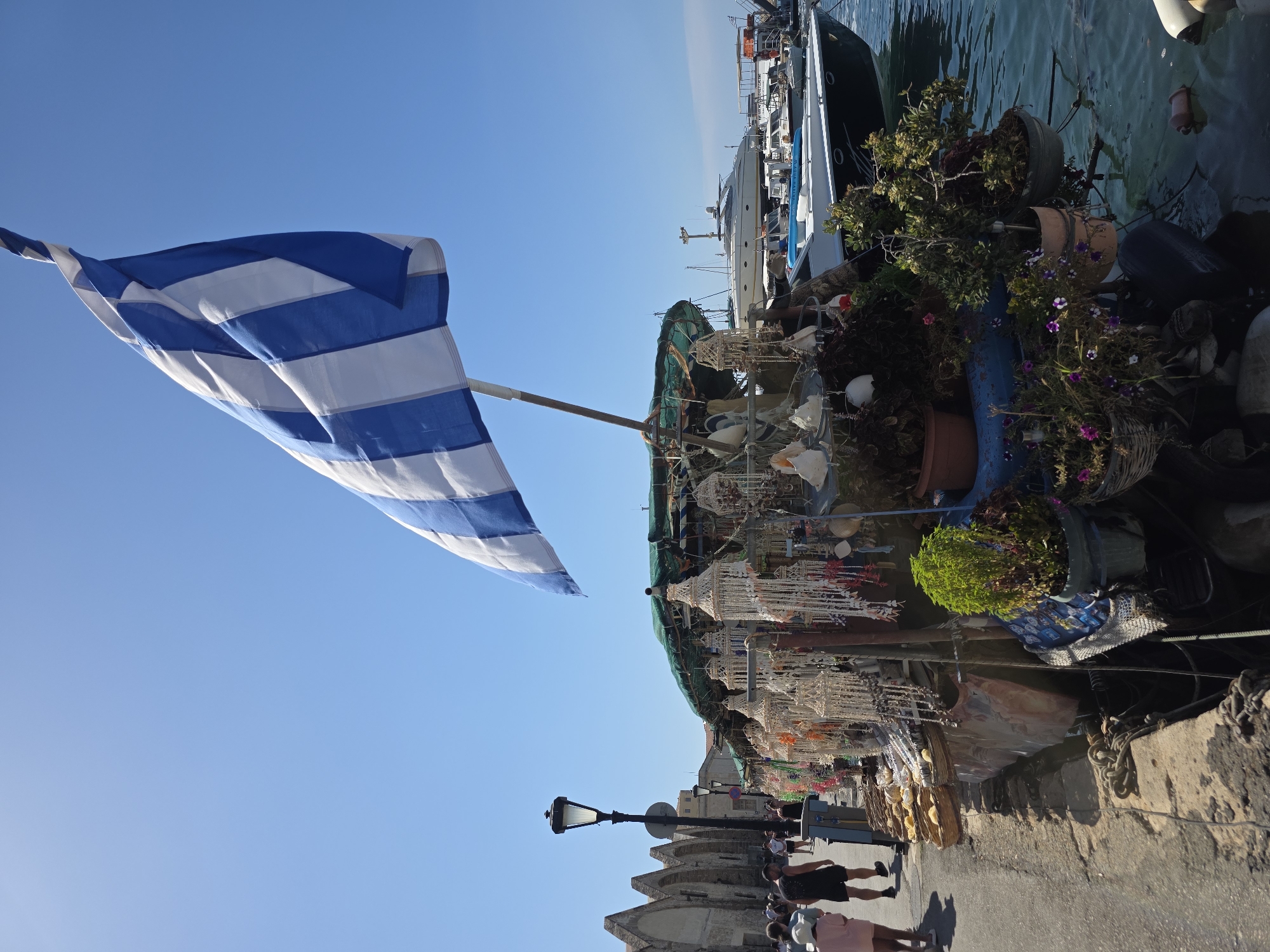 This is a medium shot, eye-level, outdoor photograph taken on a sunny day in Chania, Greece. A Greek flag is prominently displayed, waving from a pole on the right side of the frame. In the foreground, a market stall is set up along a waterfront promenade. The stall is filled with various handcrafted items, including macrame hangings, shells, and other decorative objects. Potted plants, some with flowers, are also visible at the stall.
To the left of the market stall, a paved walkway runs along the waterfront, with several people walking. One person, wearing a pink top and white hat, is walking away from the camera, while another person, wearing a black tank top and shorts, is walking towards the camera. Further down the walkway, more people can be seen. In the background, on the left, are old stone buildings with arched windows. On the right, a harbor is visible with several boats docked, including a large white yacht and a speed boat. The water in the harbor is clear and blue. The sky is a clear, bright blue. The overall scene depicts a bustling waterfront market in a historic Greek city.