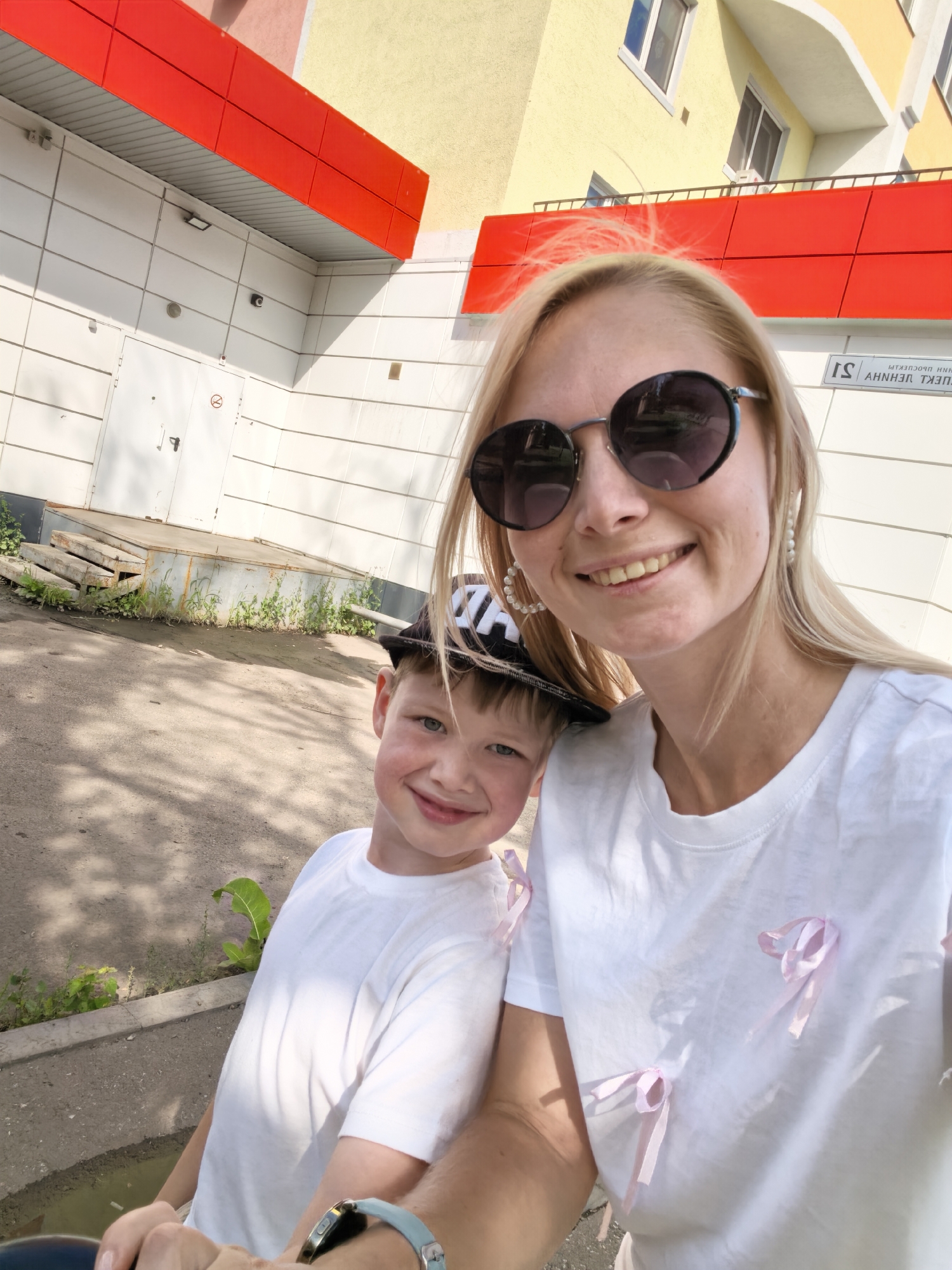 A blonde woman wearing sunglasses and a white t-shirt adorned with pink ribbons is taking a selfie with a young boy. The boy, wearing a white t-shirt and a black cap, is smiling at the camera. They are standing in front of a modern building with red and yellow accents. In the background, a street sign with Russian text is visible. The overall scene suggests an outdoor urban environment, possibly a residential area in Sterlitamak, Russia.