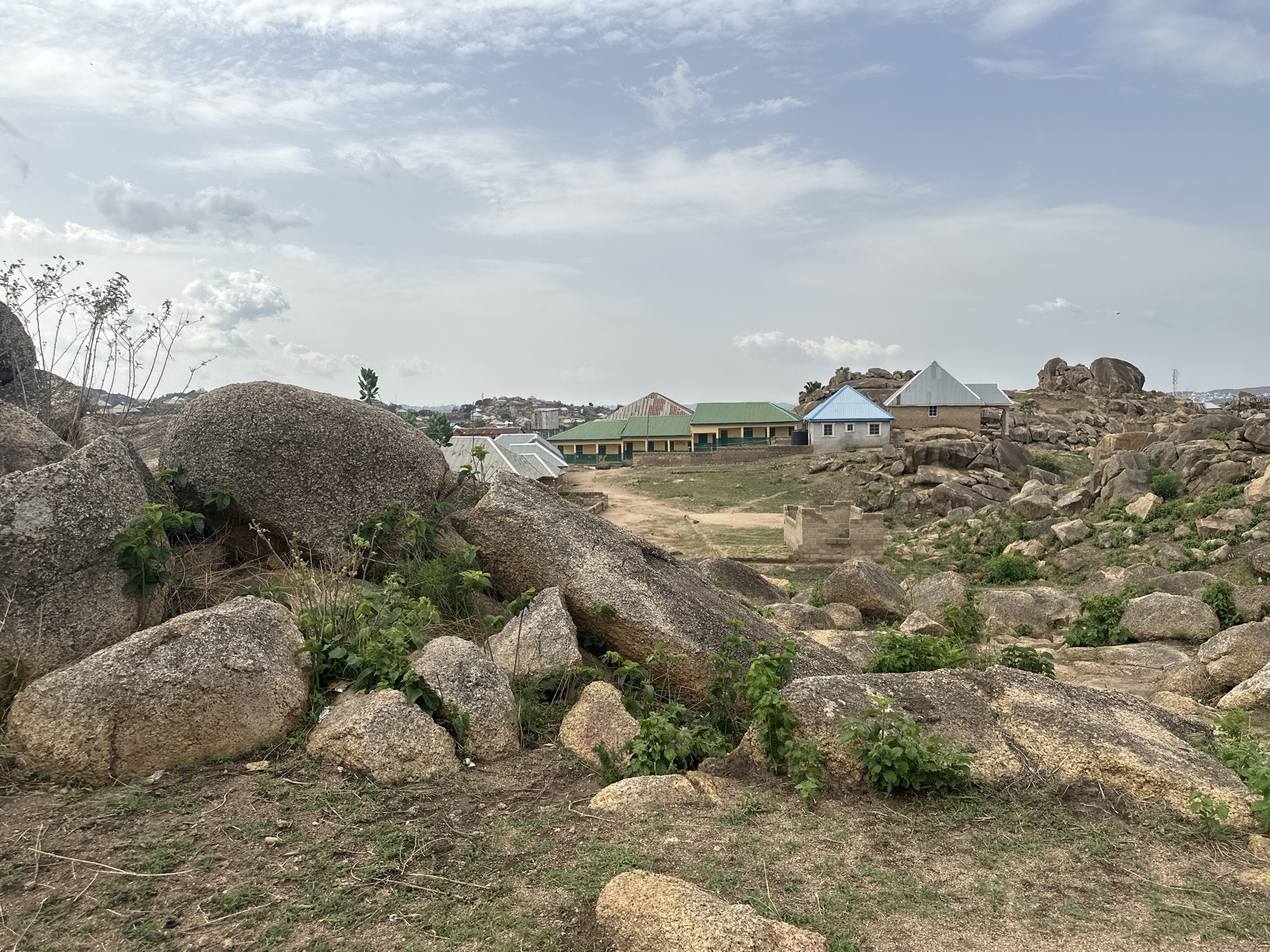 The image shows a rocky, uneven terrain in Jos, Nigeria. Large boulders and smaller rocks are scattered across the ground, with sparse vegetation growing amongst them. In the middle ground, a single-story building with a green roof and a smaller blue-roofed structure are visible. A partially constructed, square concrete structure sits to the right of the buildings. In the distance, a cityscape is faintly visible under a cloudy sky. The foreground shows dry grass and exposed soil. There are no people visible.