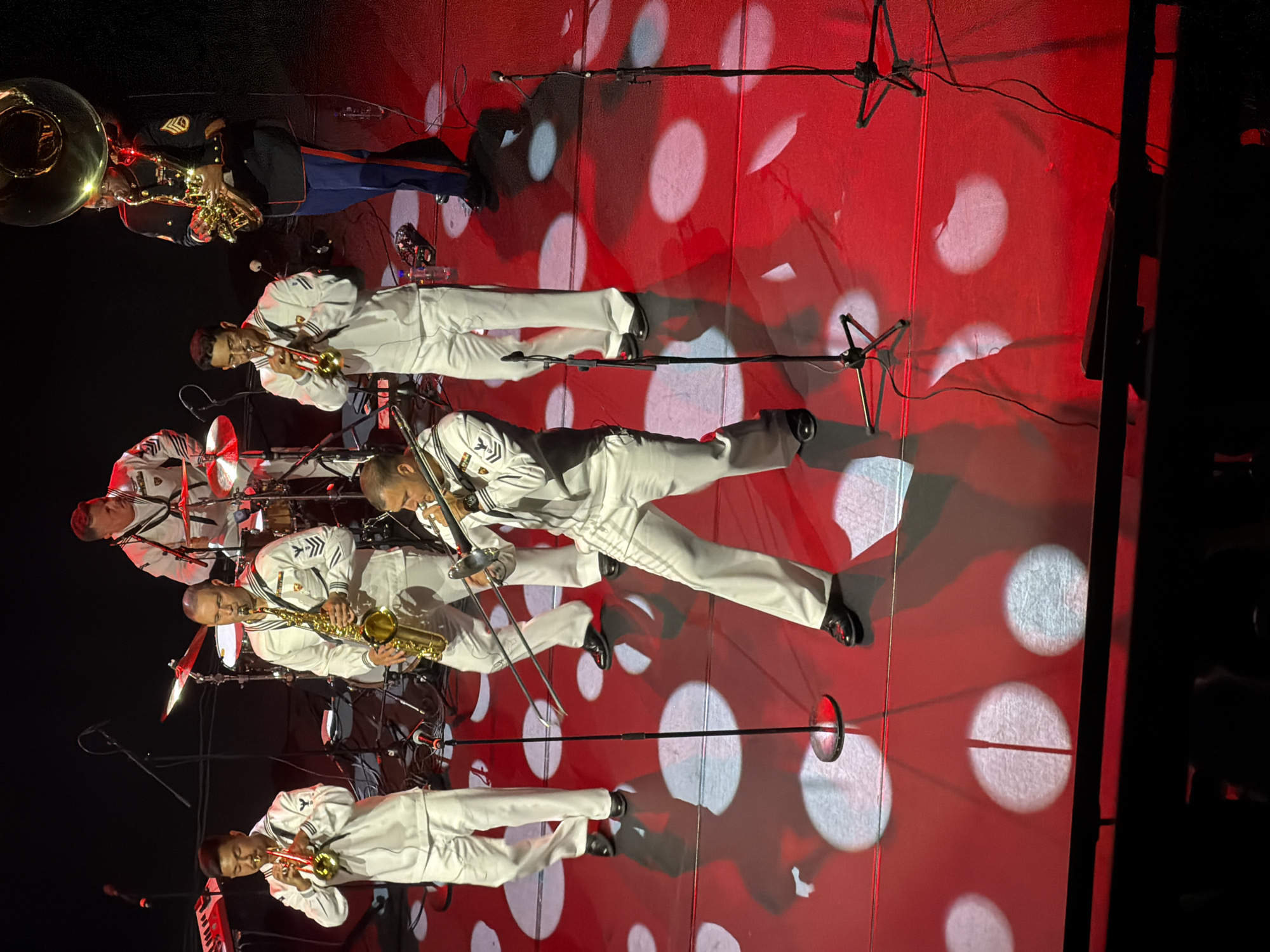 The image shows a musical performance on a stage. Several musicians are present, dressed in white naval uniforms. One musician in the foreground is playing a trombone. To his left, another musician is playing a saxophone, and further to the left, a musician plays a trumpet. In the background, another musician is seen playing drums. To the far right, a musician in a dark uniform with red and blue stripes is playing a tuba and a saxophone. The stage is lit with red lighting and circular white spotlights. The musicians are performing with various instruments, including a keyboard on the far left. The image does not provide any information about the city of Eksjö, Sweden.