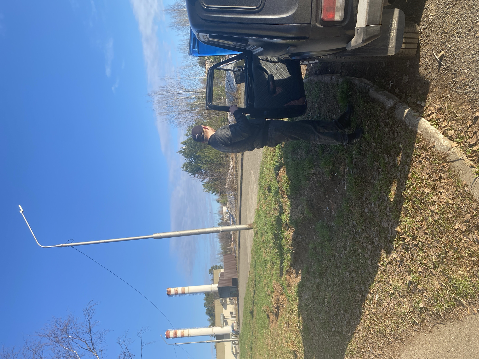 The image depicts a man standing beside a dark-colored vehicle with the driver's side door open. The man is wearing dark clothing and a baseball cap. The vehicle is parked on a patch of land covered with grass and leaves next to a curb. In the background, there is a street, trees, and a building with smokestacks. A tall metal pole with a white extension at the top is visible in the sky. The location is Марковский, Russia, as indicated by the information provided.