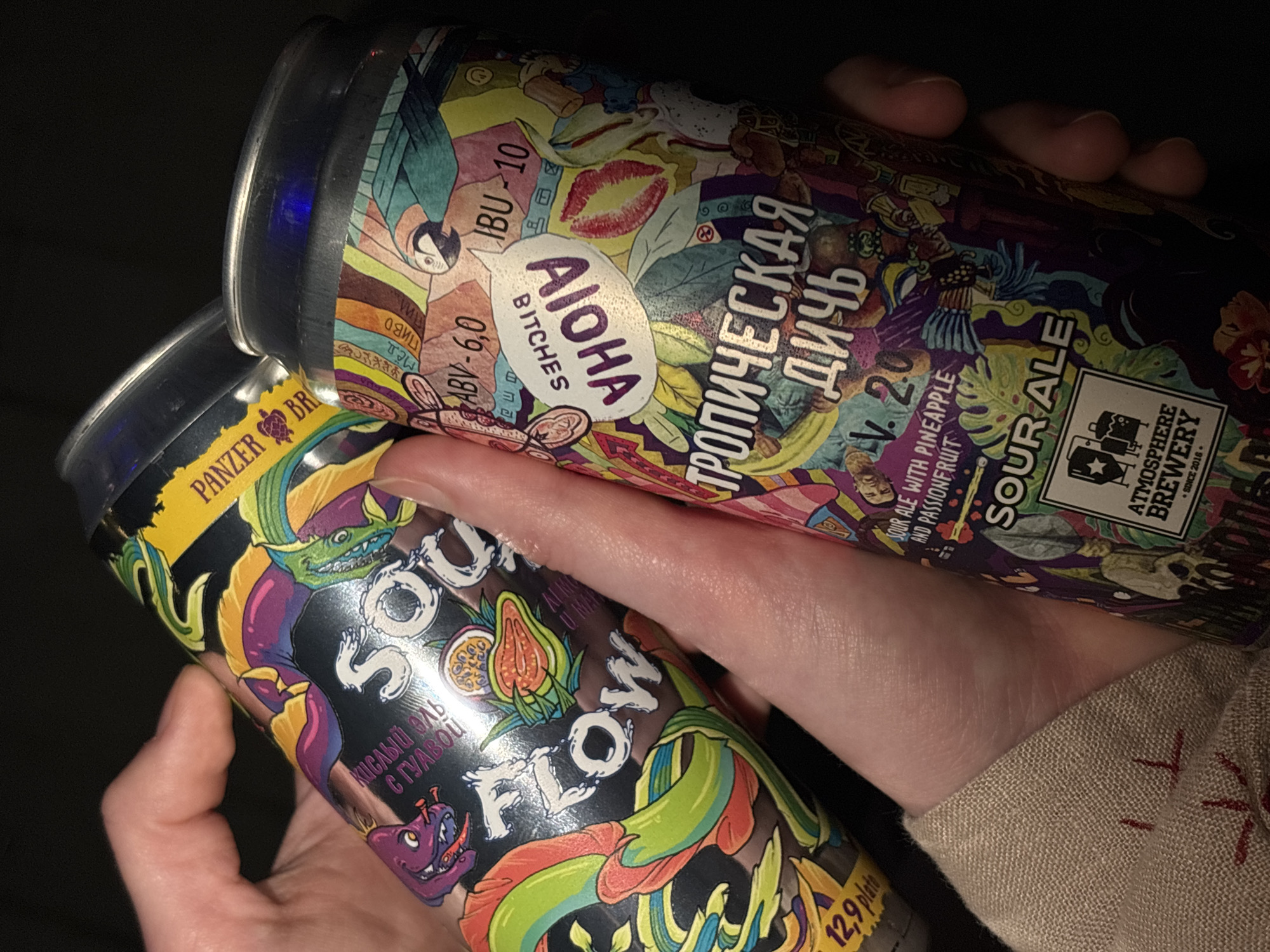 The image shows two cans of beer being held by a person. The can on the left is "Panzer Brewery Sou Flow" with a black label and colorful illustrations, with the words "Кислый эль с гуавой" (sour ale with guava) written in Russian. The can on the right is "Atmosphere Brewery Tropical Game" with a colorful and artistic label. It is a sour ale with pineapple and passionfruit, produced by Atmosphere Brewery since 2016. The label contains the words "Тропическая дичь" (tropical game) in Russian. The label also includes "Aloha Bitches". Based on the breweries named, it is likely that the location is in Perm, Russia.