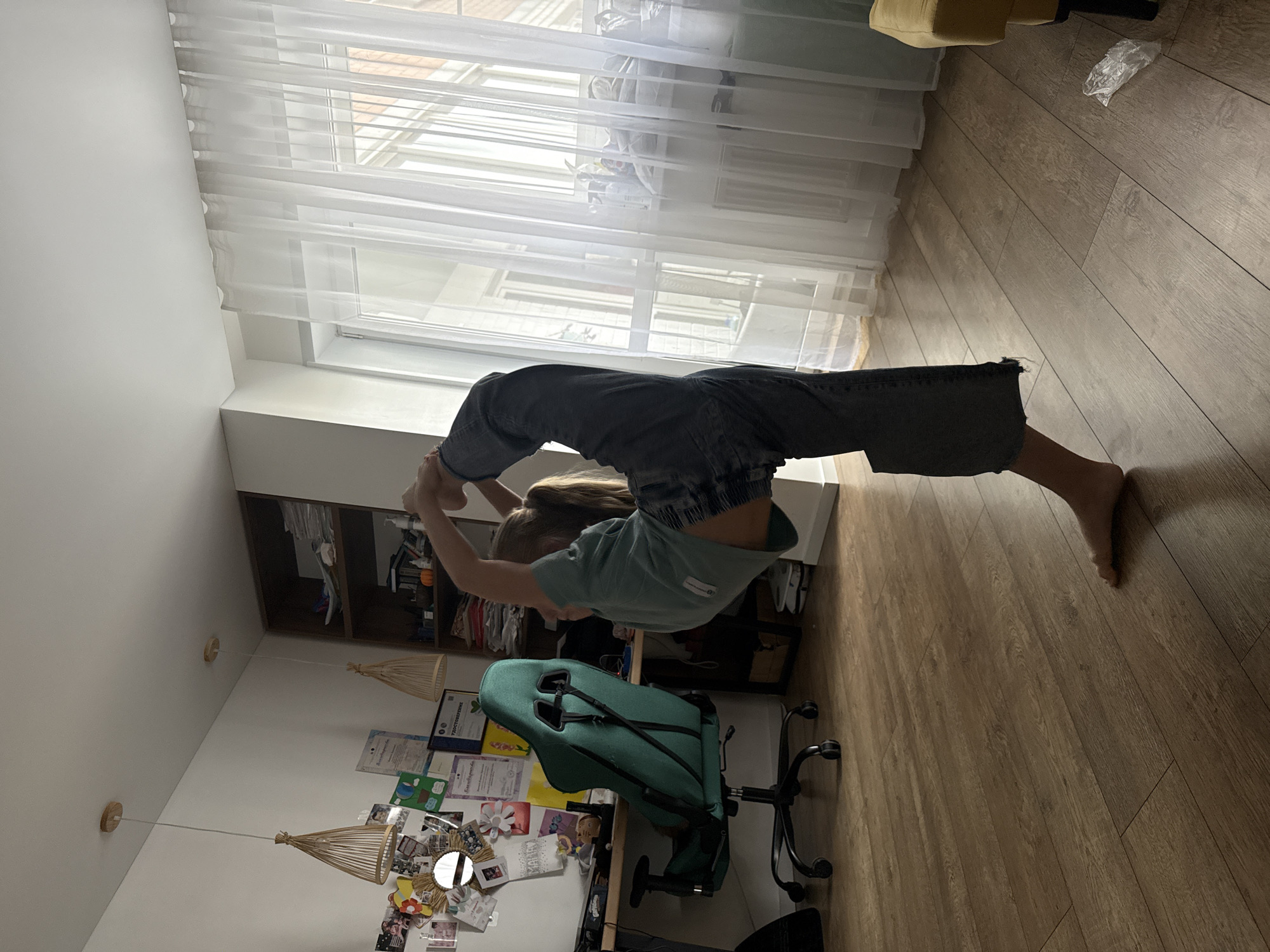 A young person is engaged in a contortionist pose inside a residence. The person is bending backwards, with their feet held by their hands, forming a complete arc. The room includes laminate flooring, a window with sheer white curtains, a desk with a green office chair, and ceiling lights. A bookshelf is visible in the background. The city of Veliky Novgorod, Russia cannot be confirmed based on the content of this image.
