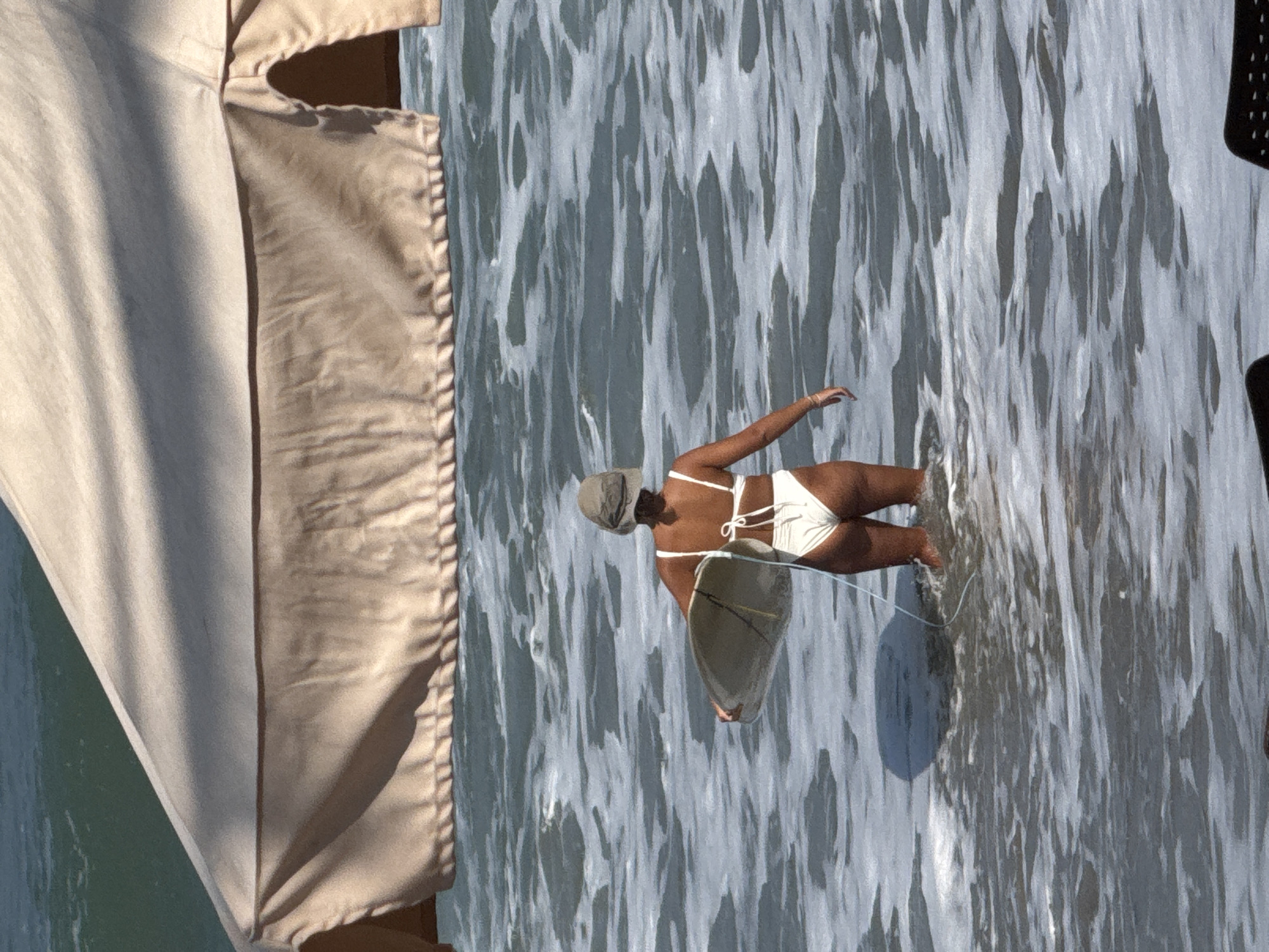 A person stands in the ocean in Kuta, Indonesia, holding a surfboard. The person is wearing a white bikini and a hat. The ocean is wavy and there is a tan umbrella above the person.