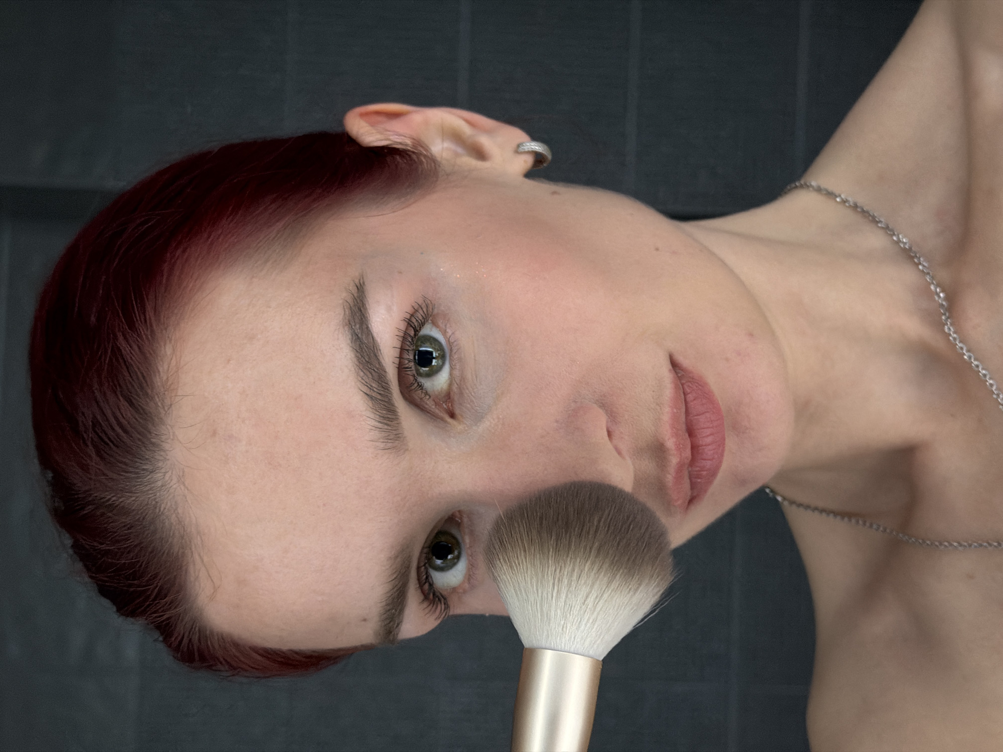 An intimate, close-up shot features a person, likely female, with fair skin, short dark red hair, and striking green eyes. The image is taken from an unusual angle, with the subject's head tilted significantly, making it appear as if they are looking upwards or have their head turned to the side, almost inverted relative to a standard portrait.
The person's gaze is directed towards the viewer. Their eyebrows are well-defined, and long, dark eyelashes frame their eyes. There is a subtle hint of blush or shimmer on their cheekbone. Their lips are slightly parted, revealing a soft pink or neutral lipstick. A small silver-toned ear cuff or hoop earring is visible on the upper part of their right ear. Around their neck, a thin, silver-toned chain necklace is visible, extending down to the collarbone area.
A fluffy makeup brush, with soft white bristles transitioning to light brown tips and a metallic gold-colored handle, is held close to the person's face, near the nose and cheek. This suggests the activity of applying makeup, possibly powder or blush.
The background is uniformly dark and appears to be a solid wall, possibly tiled or textured, creating a strong contrast with the light skin tones and the brush. The lighting is soft and even, highlighting the features of the face. No visible text is present in the image. The overall mood is focused and serene. The location context provided is Chaikovsky, Russia.