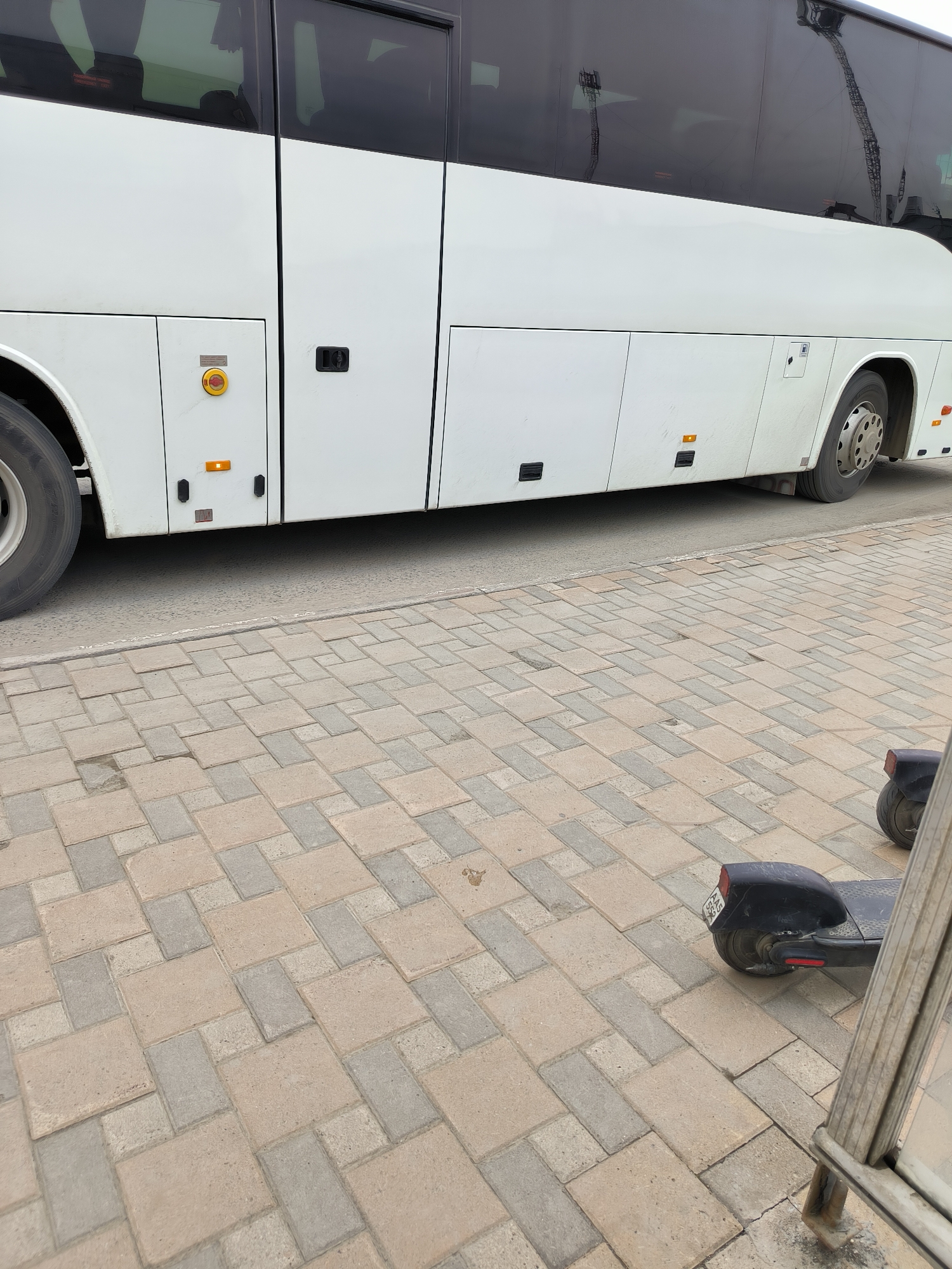 The image shows a white bus parked on the side of a street in Yuzhno-Sakhalinsk, Russia. The bus is a passenger bus with tinted windows. In front of the bus, there is a sidewalk paved with interlocking concrete blocks. Two electric scooters are parked on the sidewalk, one of which is partially visible in the lower right corner of the image. The scooters are dark-colored and appear to be for public rental. The environment appears to be an urban setting, with reflections of cranes visible in the bus windows, suggesting nearby construction or industrial activity. The lighting conditions indicate it is daytime.