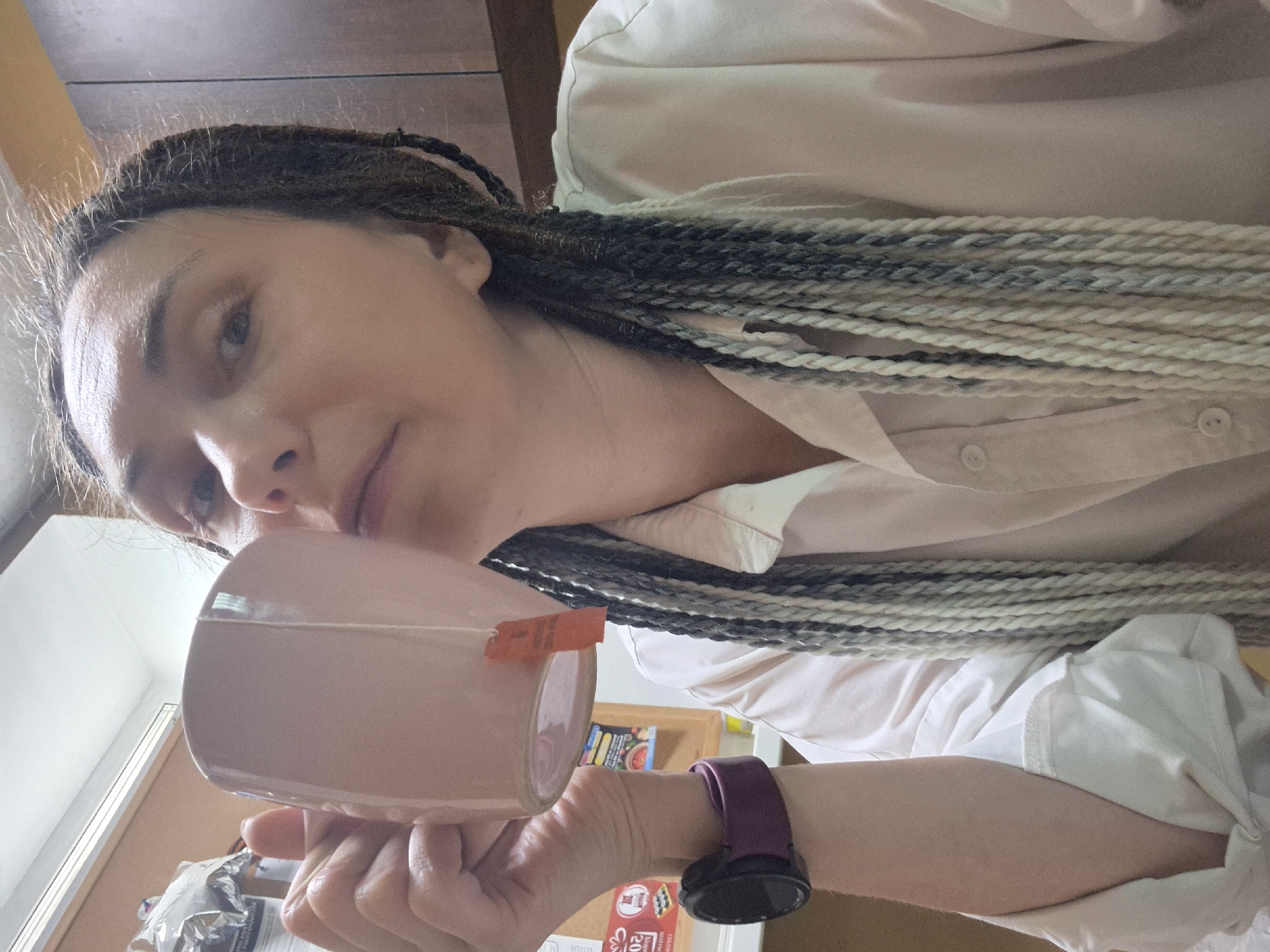 An individual with braided hair, part of which is dyed white and gray, holds a pink mug up to their mouth with their right hand. The person is wearing a light-colored collared shirt and a dark smartwatch on their left wrist. The background appears to be an indoor setting, possibly an office or kitchen, with wooden cabinets and a bulletin board visible. The lighting is moderate, suggesting an interior space with artificial light. The image is framed as a close-up shot, focusing on the person's face and upper body.
