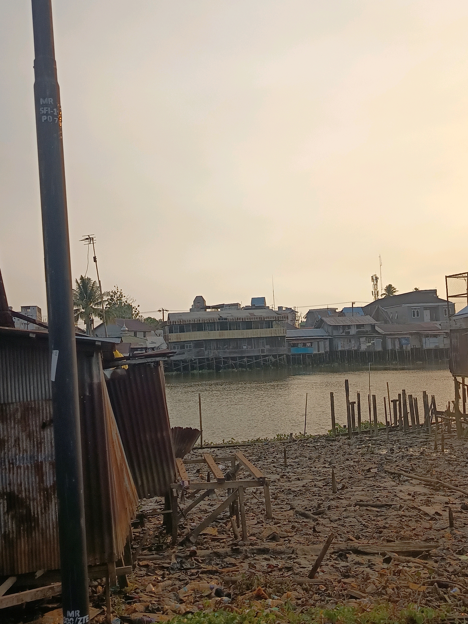 The image depicts a scene along the Martapura River in Banjarmasin, Indonesia, characterized by a densely populated urban riverside environment. In the foreground, a dark, cylindrical pole stands prominently, bearing markings that appear to be "MR SFI-1 PO7" and "MR 200/ZTE". To the left of the pole is a dilapidated structure made of corrugated metal, exhibiting signs of rust and damage. The ground in the foreground is cluttered with debris, including plastic bottles, wood fragments, and other refuse, suggesting a significant waste management issue.
The river itself occupies the midground, with its water reflecting the muted, hazy sky of what appears to be either sunrise or sunset. Along the riverbank, a series of wooden pilings jut out of the water, some intact and others broken, hinting at former structures or a current rudimentary waterfront. On the opposite bank of the river, a row of houses and buildings stands on stilts, a common architectural feature in Banjarmasin due to the city's riverine geography. These structures vary in design and condition, some appearing more modern with tiled roofs and multiple stories, while others are simpler and weathered. Visible structures include what looks like an older, longer building with a colonnade, and various residential houses. An antenna is visible on one of the rooftops, and a palm tree sprouts behind some of the structures. The overall atmosphere is one of a lived-in, bustling, yet somewhat neglected urban riverside community.