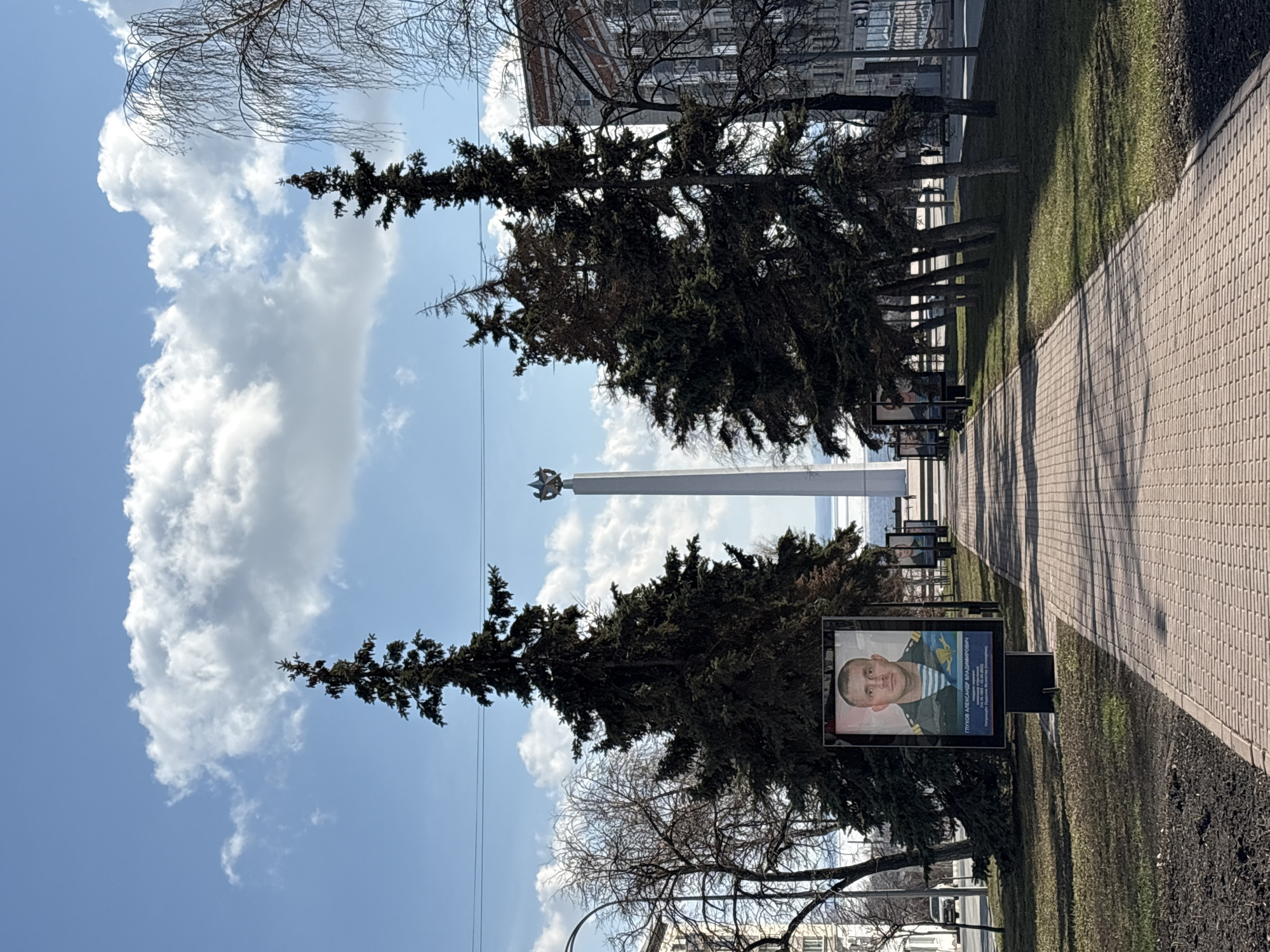 The image depicts a memorial pathway in Ulyanovsk, Russia. A brick pathway runs along a green strip, bordered by trees. Along the pathway are portrait displays honoring military personnel. A tall, white obelisk topped with a star is visible in the background. The sky is blue with scattered clouds. Buildings are visible in the distance, indicating an urban setting.