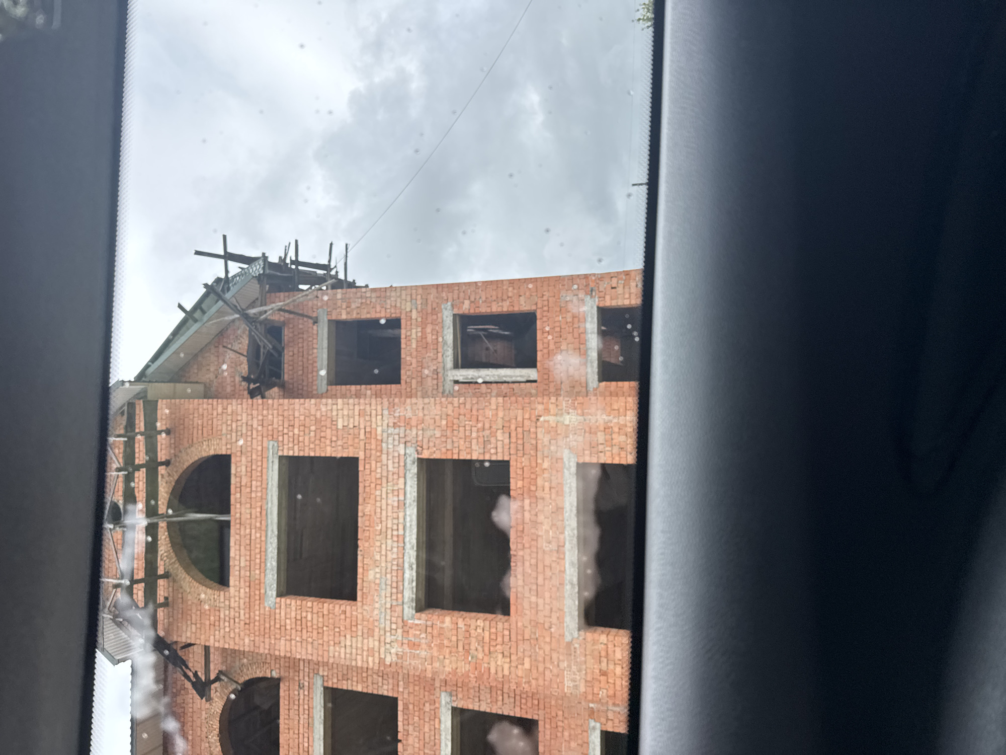 An unfinished brick building is viewed through a car windshield. The building is multi-story with numerous rectangular openings for windows. Scaffolding is visible on the upper floors and roofline. The sky is overcast with clouds. The foreground is dominated by the dark interior of the car, obscuring the lower part of the scene. There are water droplets on the windshield.