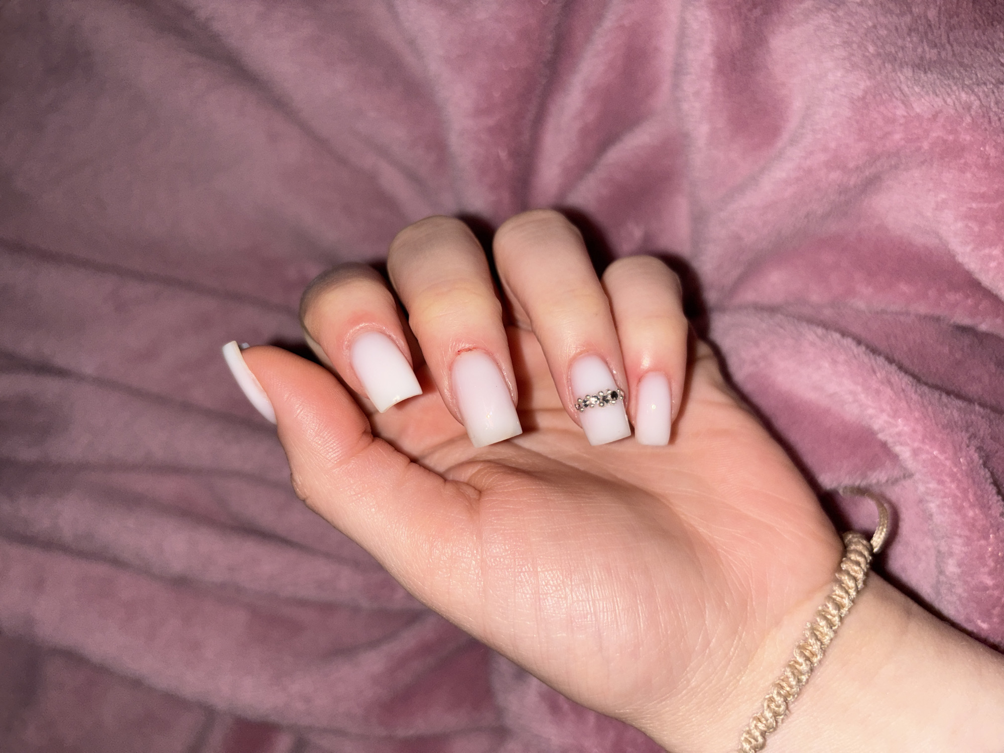 A close-up shows a fair-skinned human hand with manicured, square-shaped, milky-white nails resting against a soft, plush, pink fabric. A silver band with embedded crystals or stones adorns the middle finger of the hand. A woven bracelet with a light beige or tan color is visible around the wrist. The location is Unknown, Unknown.