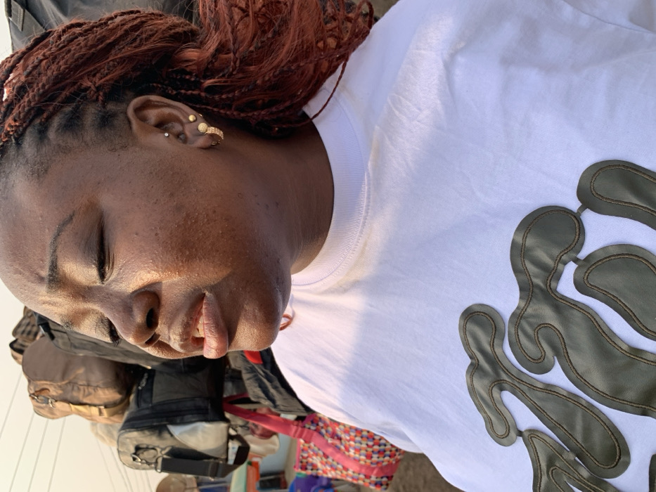 A close-up depicts a woman with reddish-brown braided hair styled in a bun. Her skin tone is dark, and she has multiple ear piercings adorned with gold jewelry. Her eyes are closed. She's wearing a white t-shirt featuring an abstract pattern or design in a muted green or olive tone. A dark-colored bag or backpack rests against her, and a brightly colored woven bag is visible in the background.