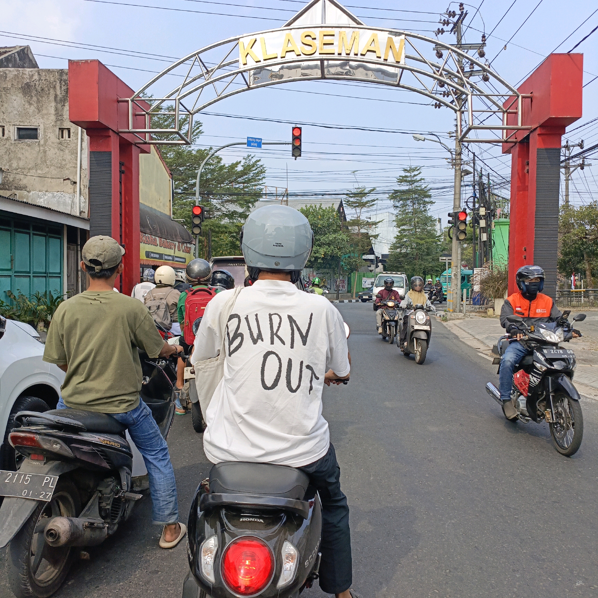 The image depicts a street scene in Salatiga, Indonesia, featuring a prominent archway with "KLASEMAN" written in gold lettering. Several motorcyclists are present, indicating traffic. One motorcyclist in the foreground wears a white t-shirt with the words "BURN OUT" printed on the back. The environment includes buildings, power lines, and traffic lights, one of which is red, suggesting that the vehicles are stopped or preparing to stop. The overall impression is a typical urban street with moderate traffic.